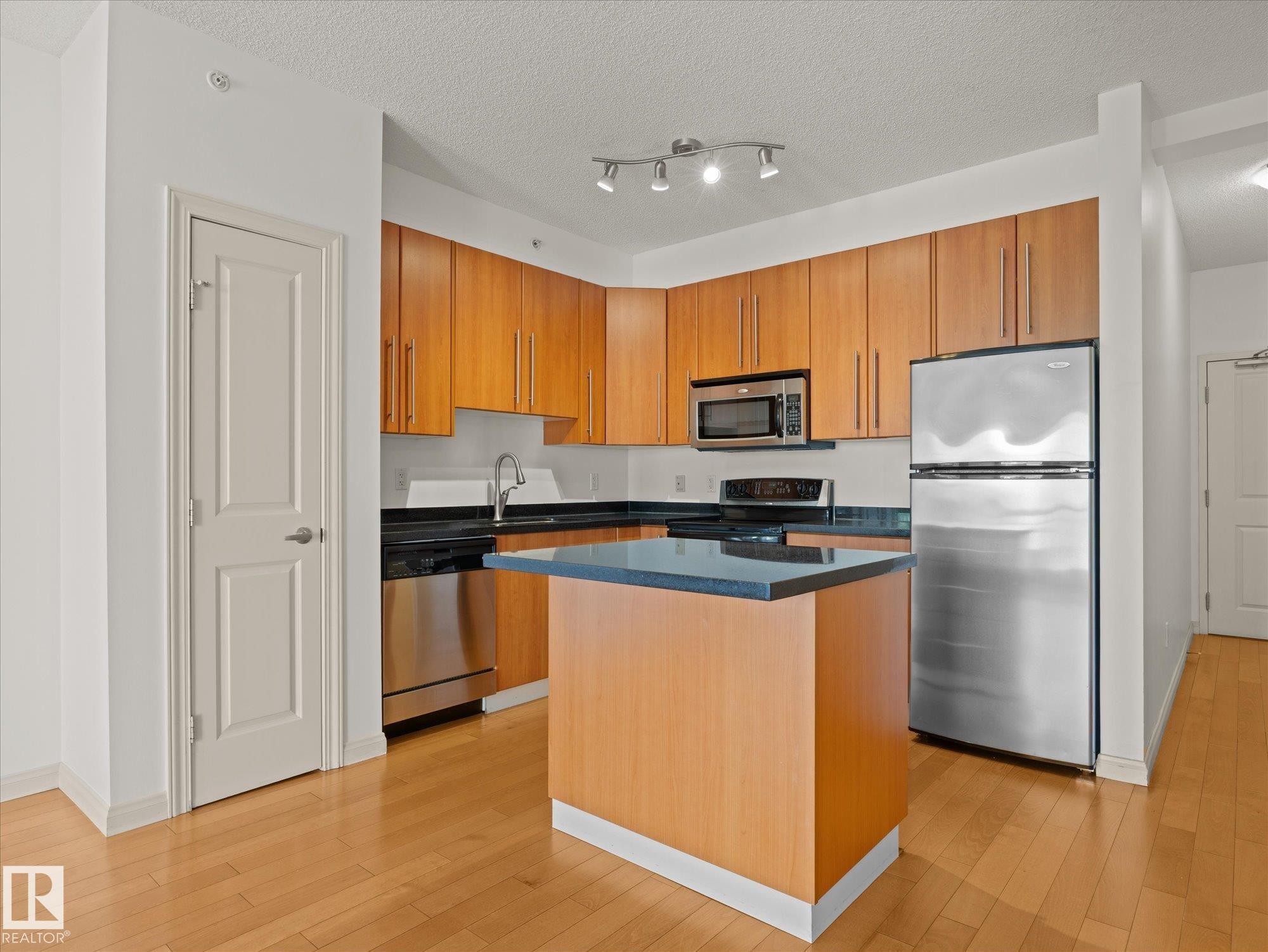 Kitchen featuring wood-finish cabinetry, stainless steel appliances, dark countertops, and a central island with a contrasting dark surface - #707 9819 104 St Nw, Edmonton, AB