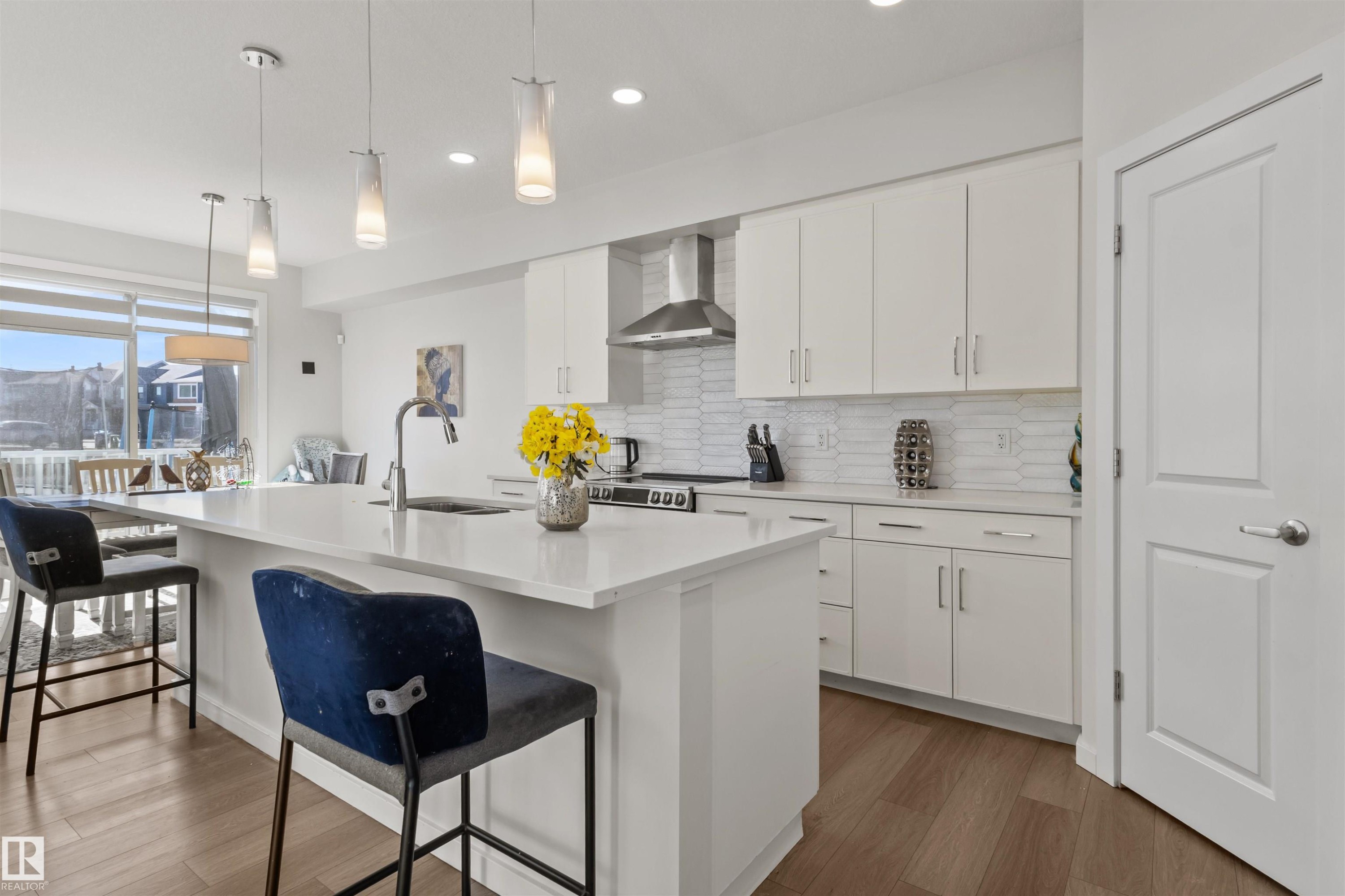 Contemporary kitchen featuring a large island with a countertop, white cabinetry, a stainless steel range hood, a tiled backsplash, and wood-finish flooring - 1471 Plum Circle Sw, Edmonton, AB - Indoor Photo Showing Kitchen With Upgraded Kitchen