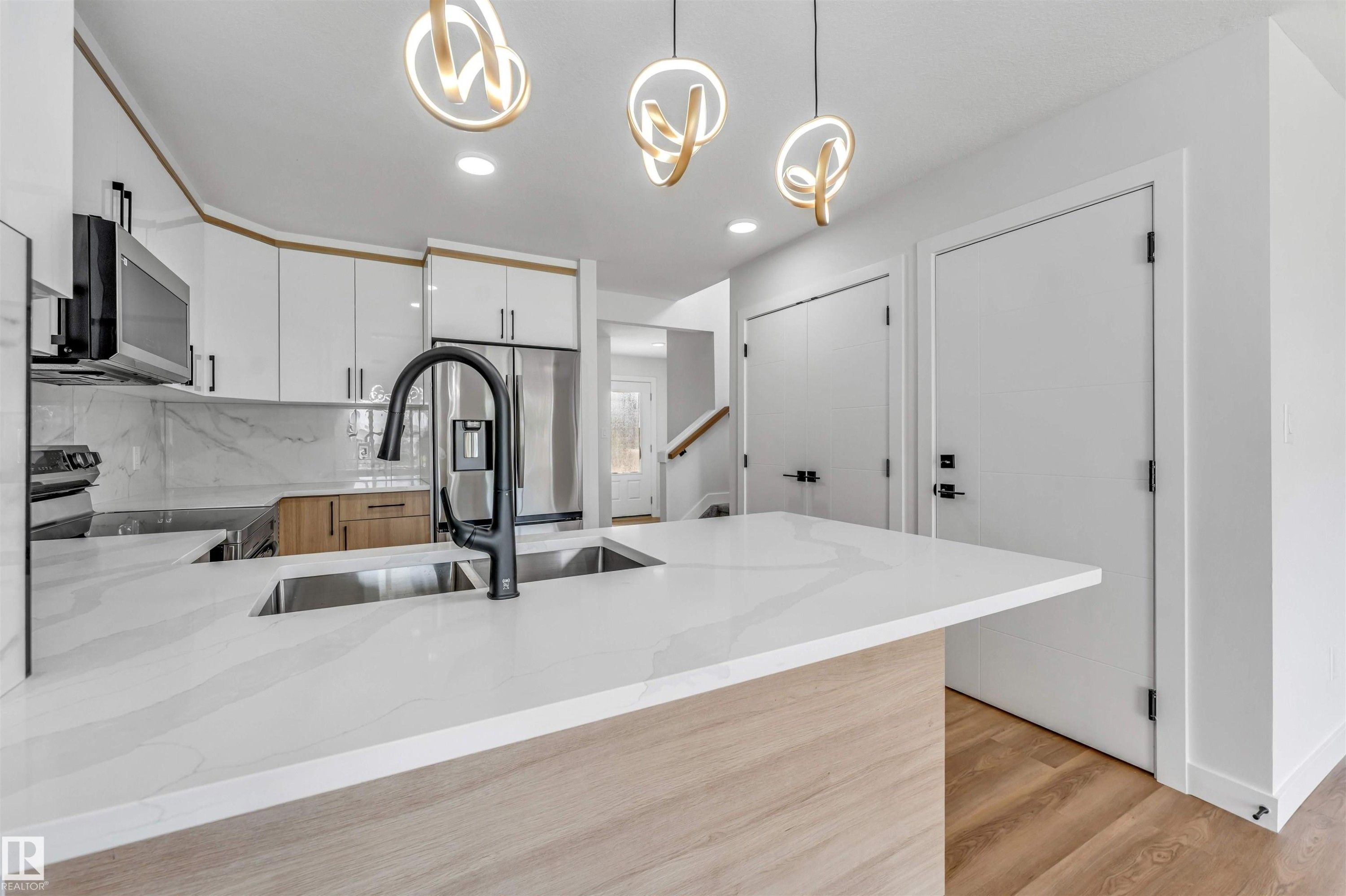 Kitchen featuring an expansive island with a double stainless steel sink and a matte black faucet, complemented by white cabinetry, stainless steel appliances, and wood-finish flooring - 1712 27 St Nw, Edmonton, AB - Indoor Photo Showing Kitchen With Double Sink With Upgraded Kitchen