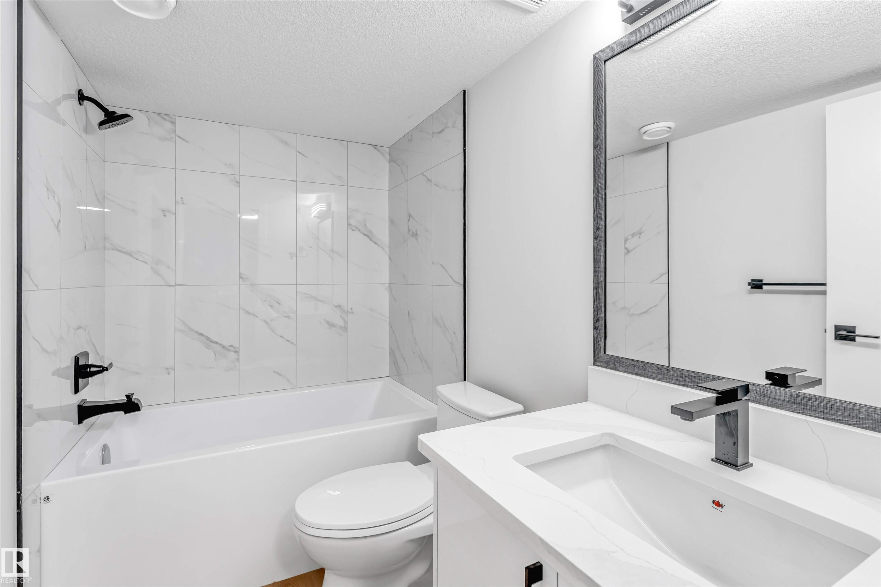 Modern bathroom featuring a white bathtub with subway tile surround, matte black shower fixtures, a toilet, a vanity with an integrated sink, and a framed mirror - 1712 27 St Nw, Edmonton, AB - Indoor Photo Showing Bathroom