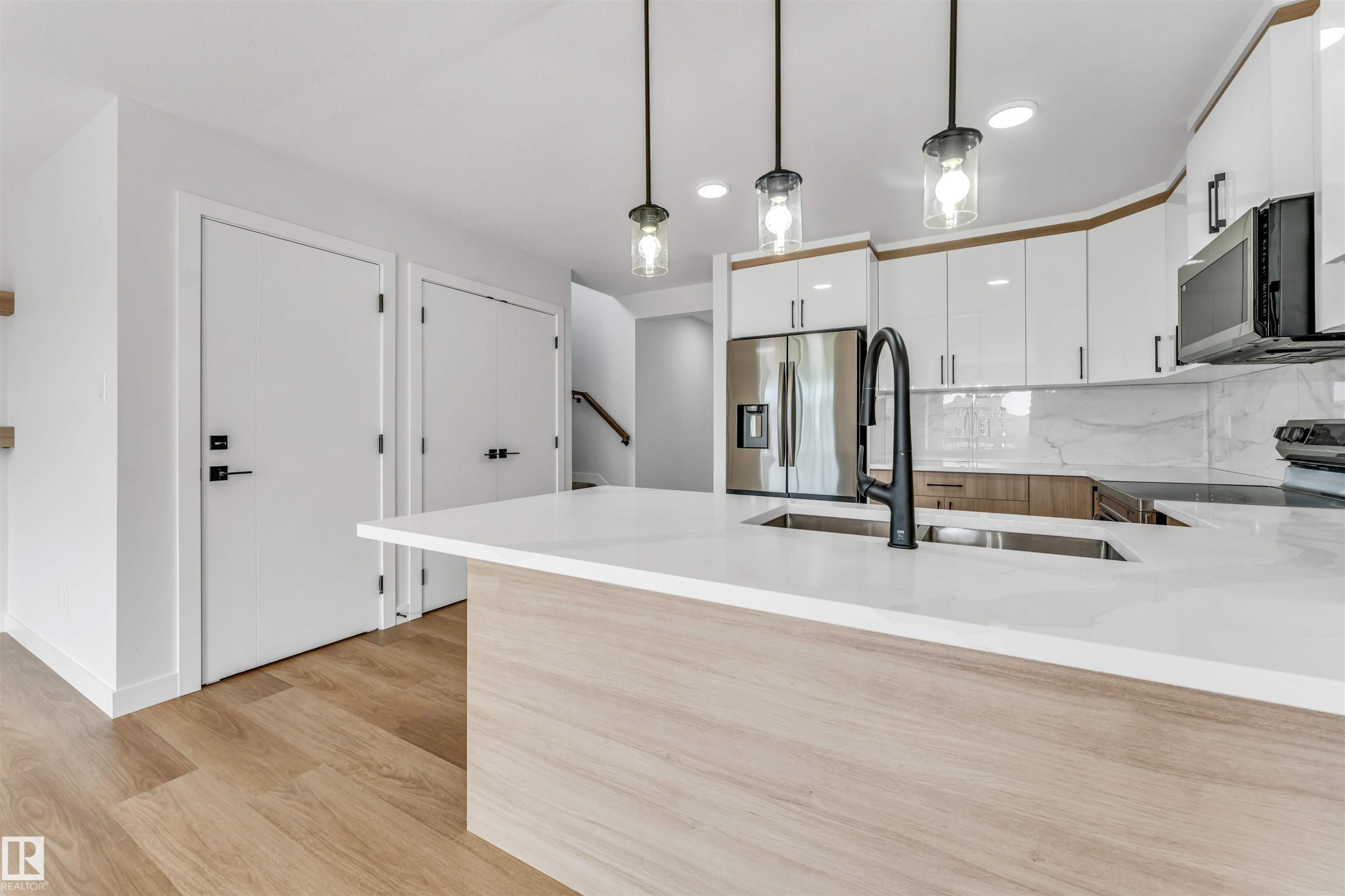 Kitchen featuring a large island with a double basin sink and matte black faucet, light wood-finish flooring, white flat-panel cabinetry, stainless steel appliances, and a marble-look backsplash - 1714 27 St. Nw, Edmonton, AB - Indoor Photo Showing Kitchen With Upgraded Kitchen