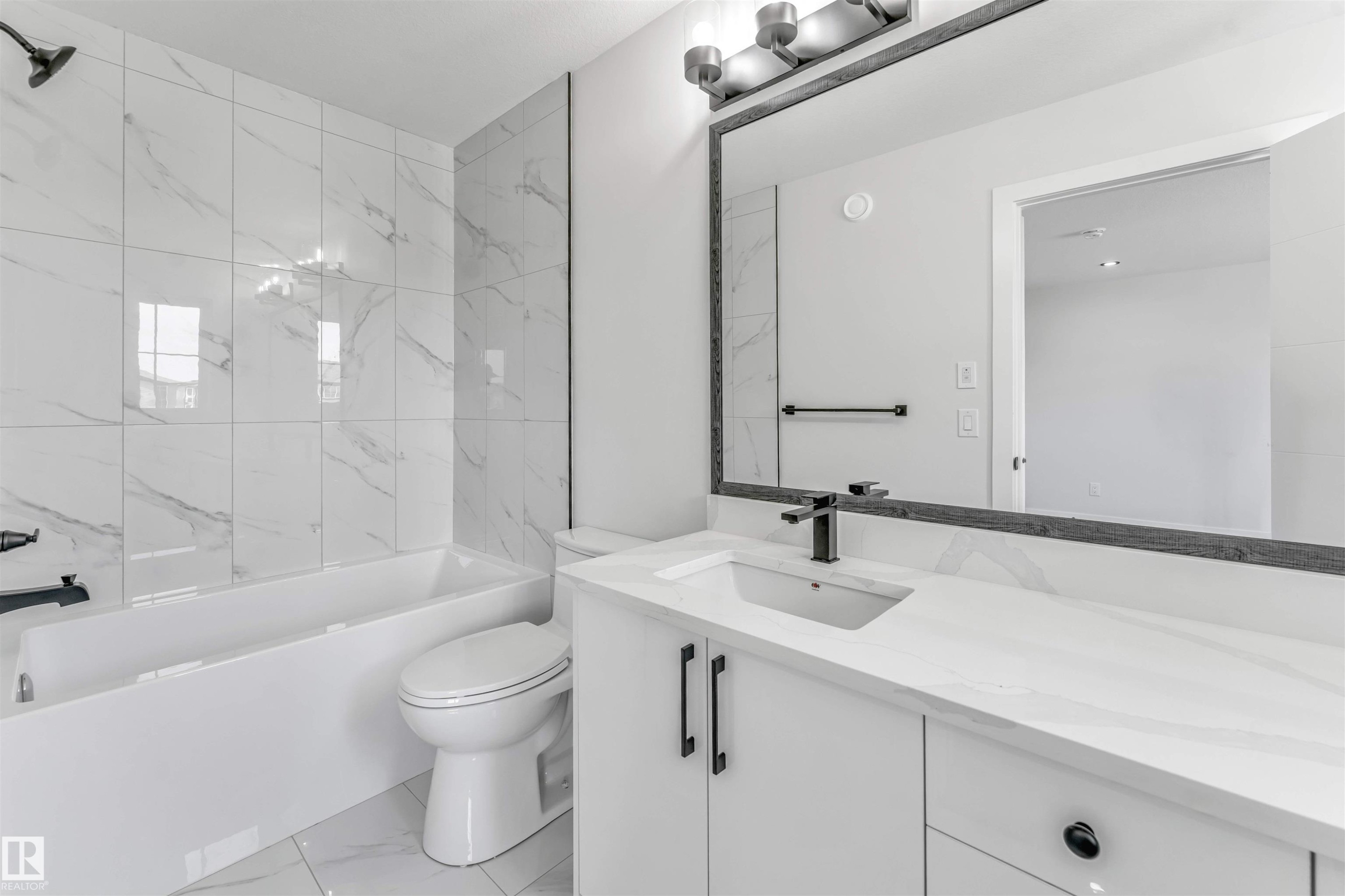 Contemporary bathroom featuring a white vanity with integrated sink, black fixtures, a framed mirror, and a built-in tub with white-veined tile surround - 1714 27 St. Nw, Edmonton, AB - Indoor Photo Showing Bathroom