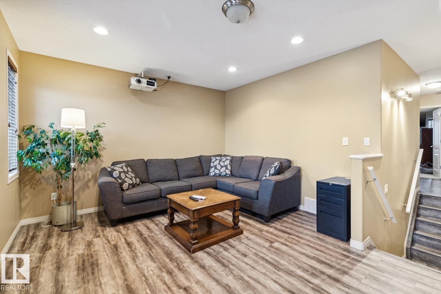 Recreation room featuring wood-finish flooring, recessed ceiling lighting, a wall-mounted projector, and a staircase with a white handrail - 16765 119 Street Nw, Edmonton, AB - Indoor