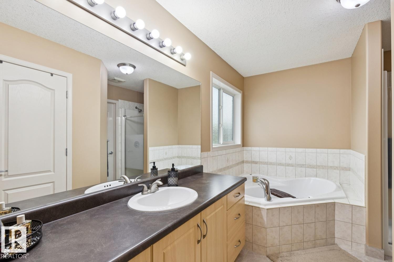 Bathroom featuring a corner soaking tub with tile surround, a single vanity with wood-finish cabinetry, a dark countertop, and a large mirror with strip lighting - 16765 119 Street Nw, Edmonton, AB - Indoor Photo Showing Bathroom