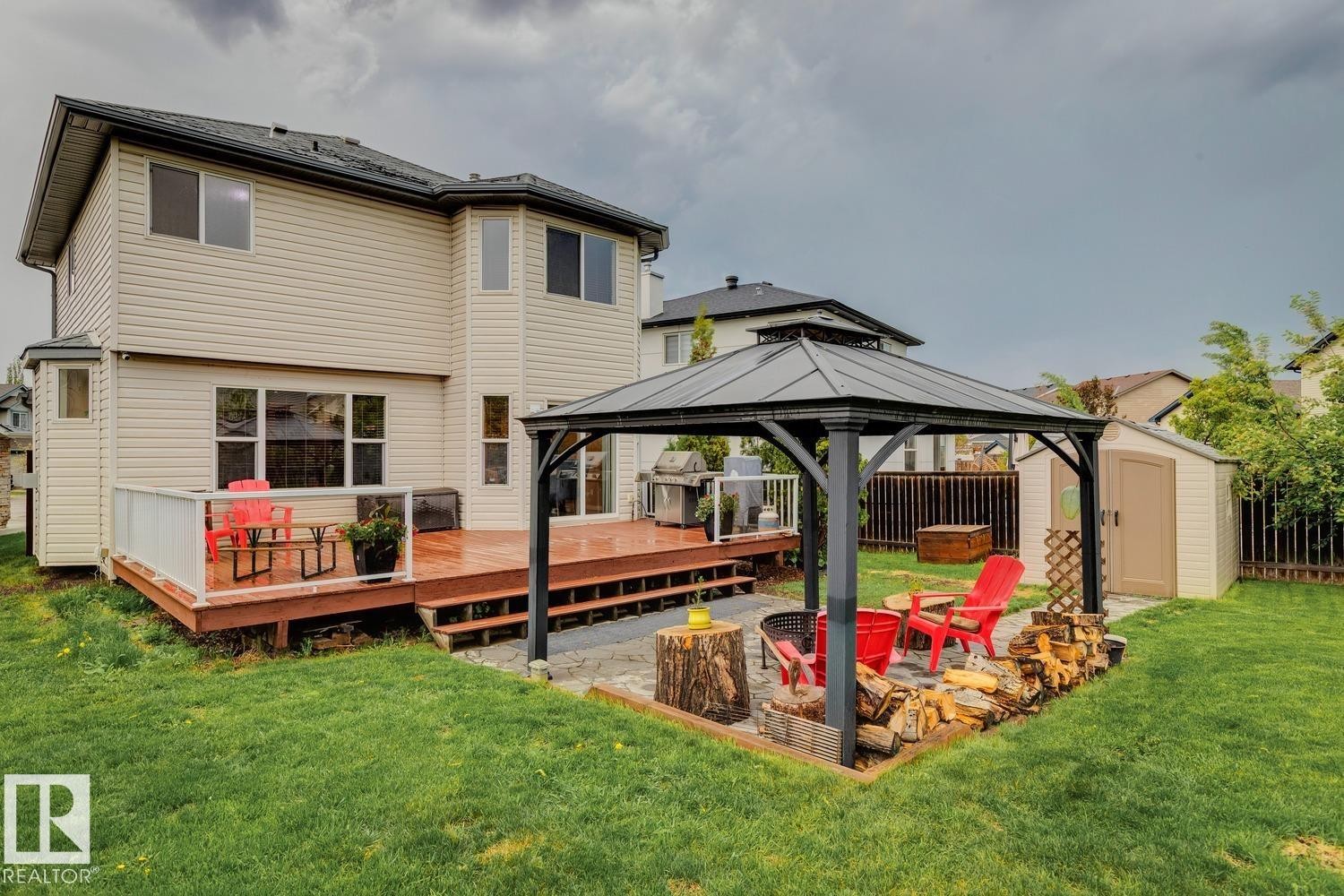 Expansive backyard featuring a wood deck with glass railing, a gazebo over a paver patio with a built-in fire pit, a storage shed, and a privacy fence - 16765 119 Street Nw, Edmonton, AB - Outdoor With Deck Patio Veranda