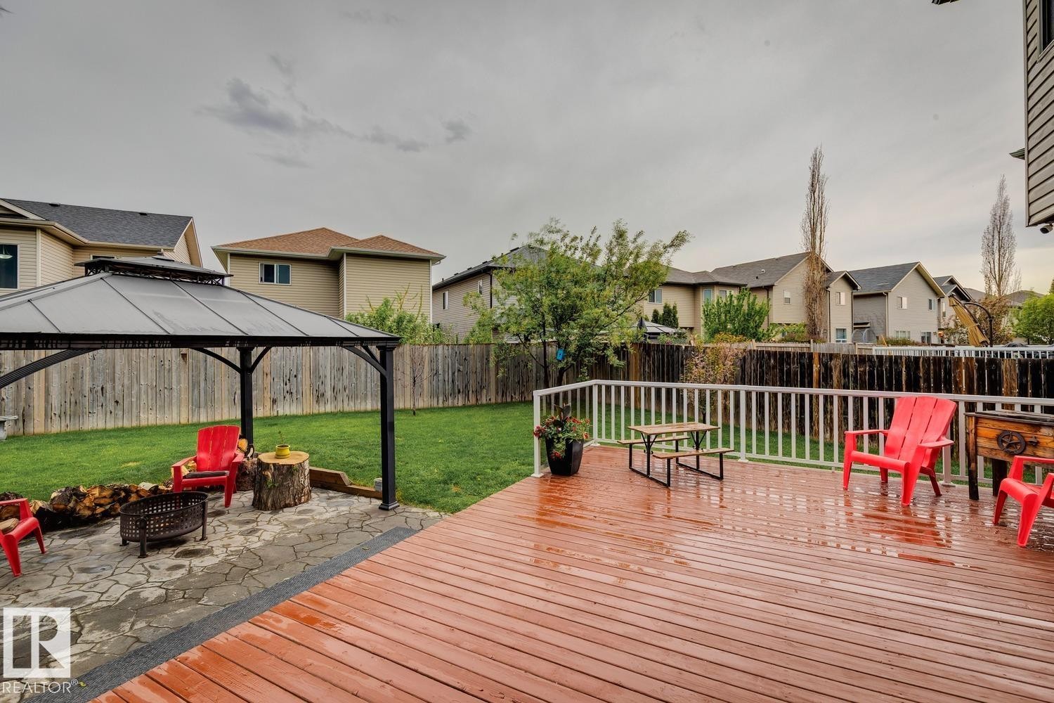Expansive backyard featuring a wood-finish deck with white railing, a stone patio with a fire pit, and a black gazebo with a hardtop roof - 16765 119 Street Nw, Edmonton, AB - Outdoor With Deck Patio Veranda With Exterior