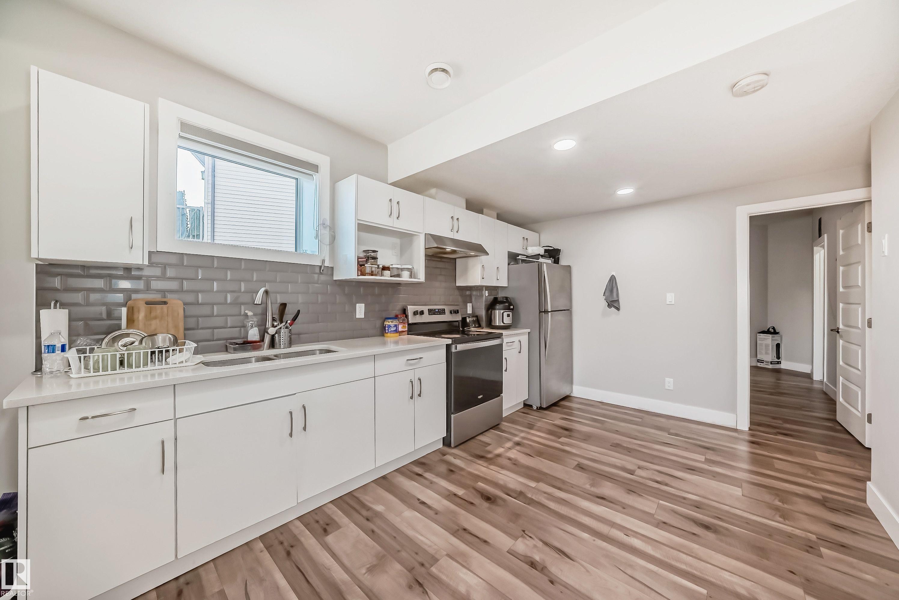 Contemporary kitchen featuring white cabinetry, a subway tile backsplash, stainless steel appliances, a window above the sink, and wood-finish flooring - 2464 15 Avenue Nw, Edmonton, AB - Indoor Photo Showing Kitchen With Double Sink