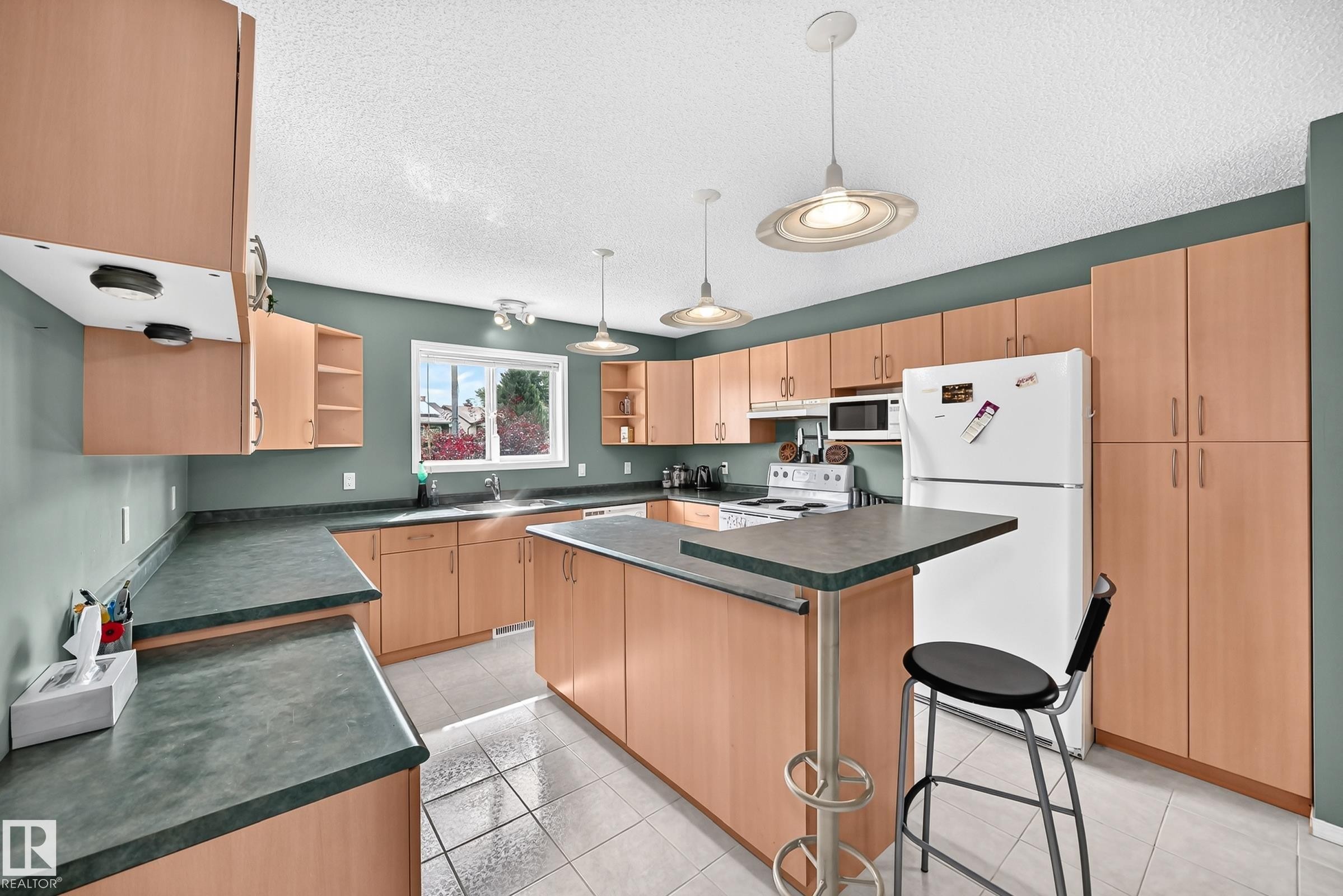 Kitchen featuring extensive light wood-finish cabinetry, dark countertops, white tile flooring, and two pendant lights - 1116 39 Street Nw, Edmonton, AB - Indoor Photo Showing Kitchen
