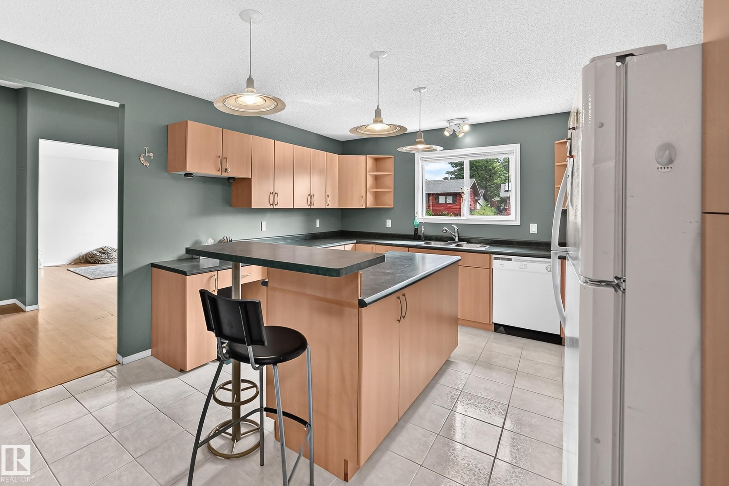 Kitchen featuring wood cabinetry, dark countertops, a central island with an extended breakfast bar, white tile flooring, and pendant lighting - 1116 39 Street Nw, Edmonton, AB - Indoor Photo Showing Kitchen