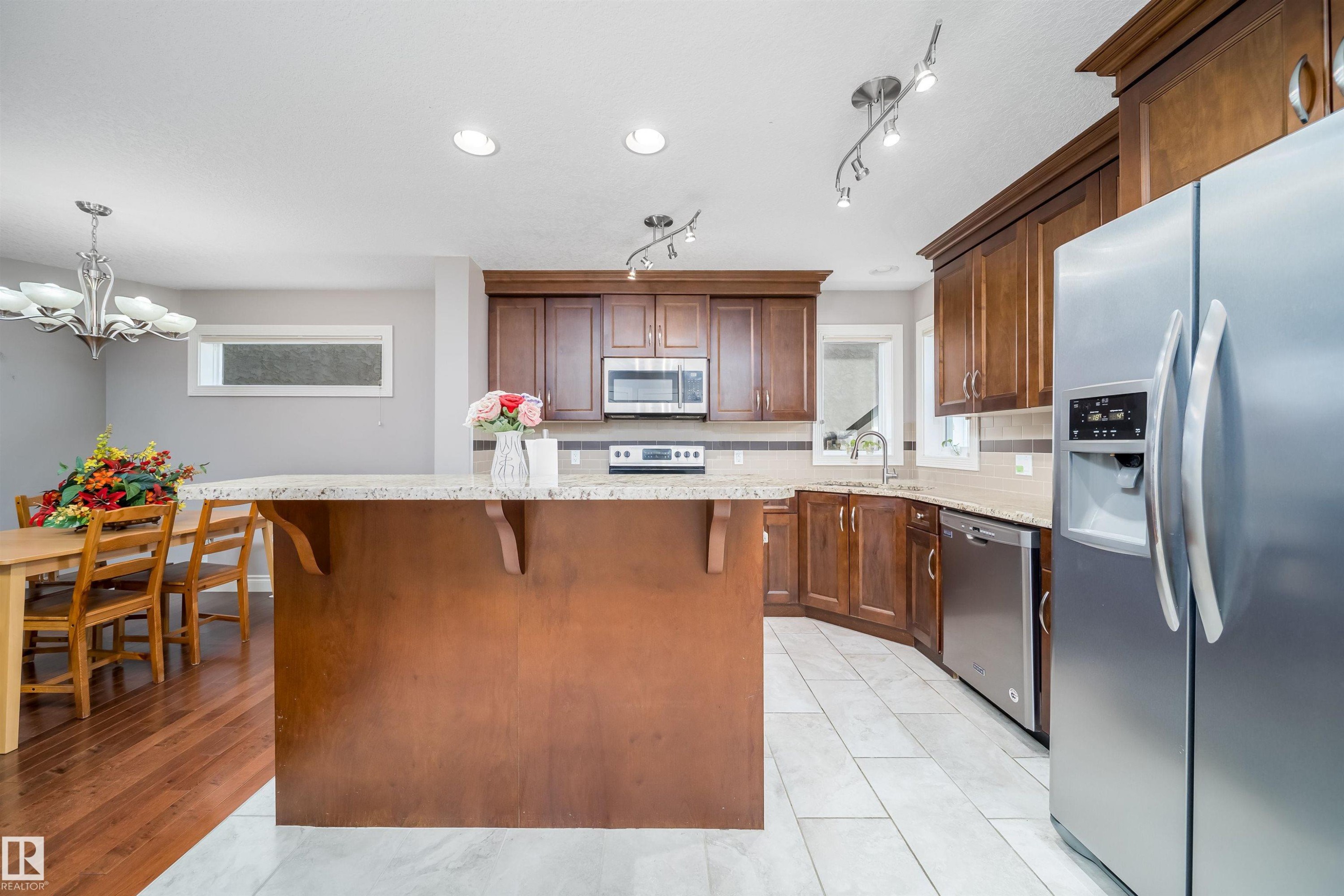 Kitchen featuring dark wood-finish cabinetry, stainless steel appliances, light-toned countertops, tile flooring, and a central island with bar seating - 11342 79 Avenue, Edmonton, AB - Indoor Photo Showing Kitchen