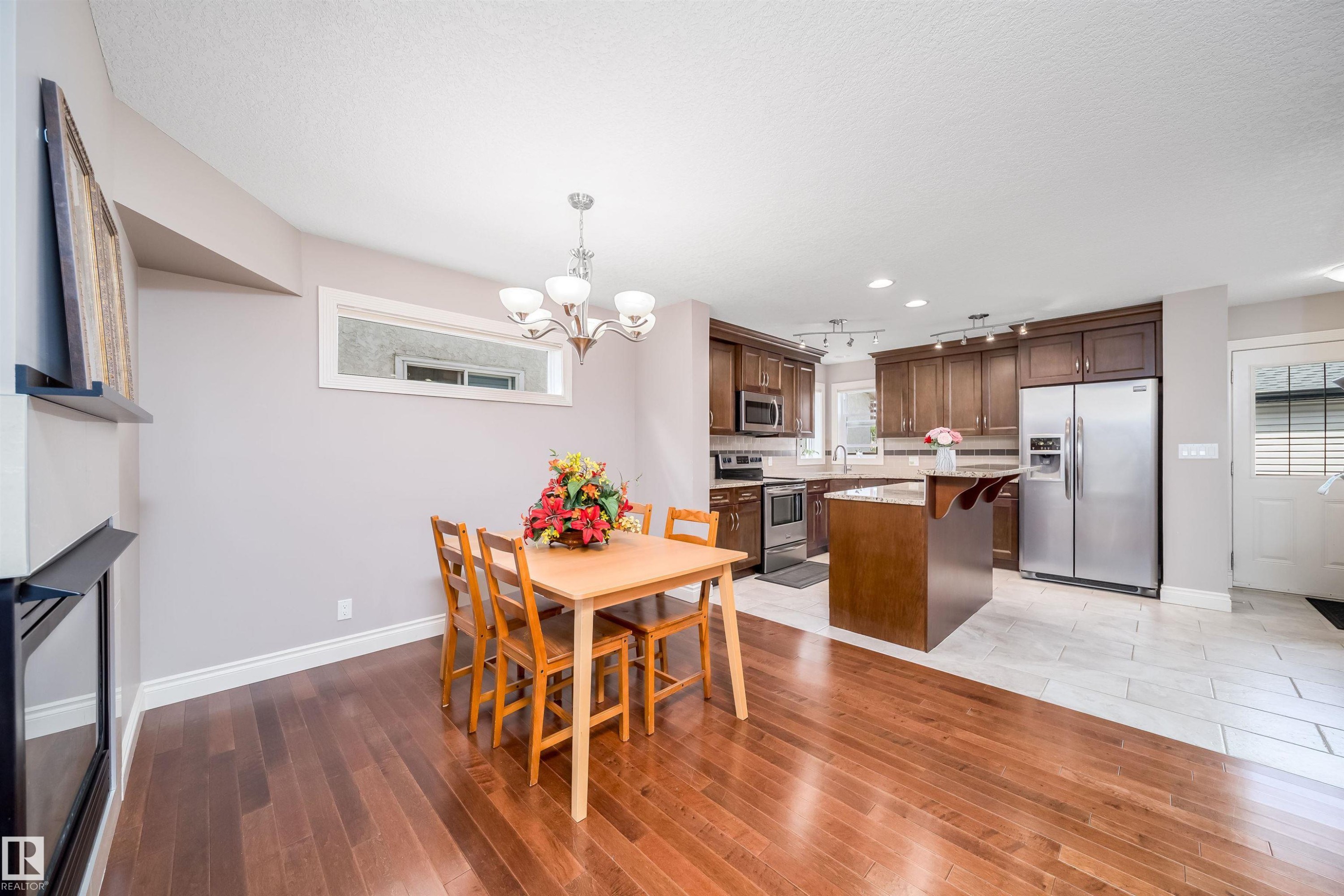 Expansive kitchen and dining area featuring wood-finish flooring, stainless steel appliances, dark wood cabinetry, a center island with countertop seating, and a built-in electric fireplace - 11342 79 Avenue, Edmonton, AB - Indoor