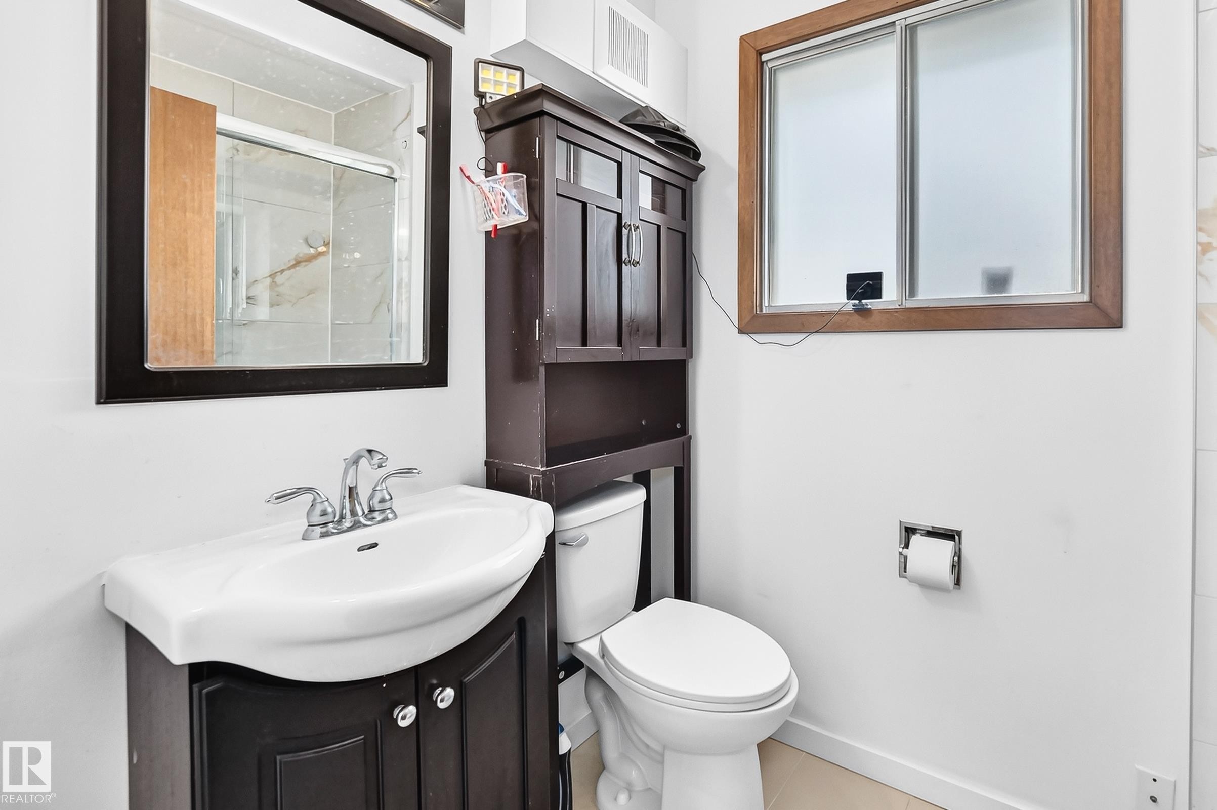 Bathroom featuring a wood-finish framed mirror, a vanity with a white ceramic sink, a chrome faucet, a toilet, and a wood-frame window with frosted glass - 1928 68 Street Nw, Edmonton, AB - Indoor Photo Showing Bathroom