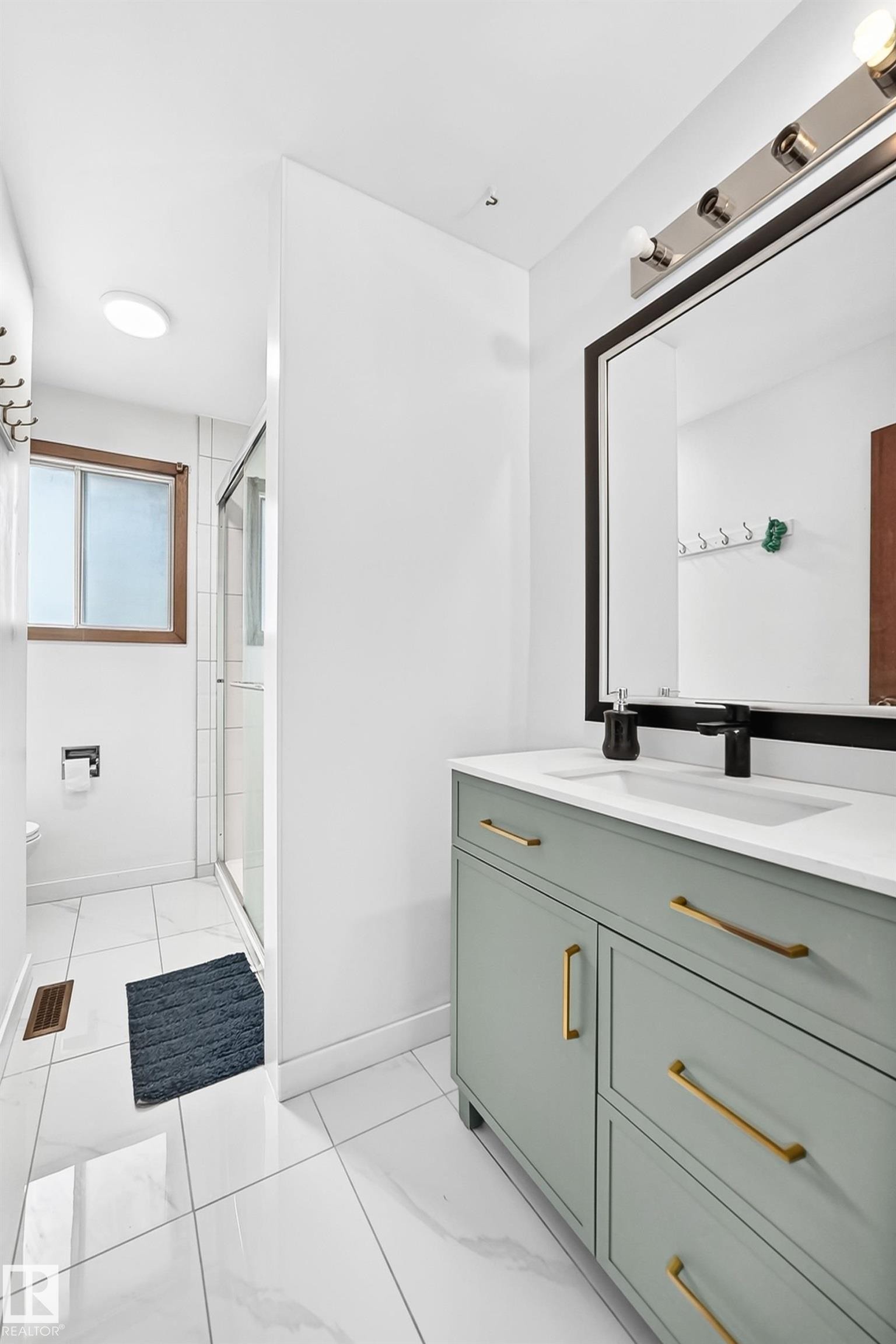 Modern bathroom vanity featuring light green cabinetry with gold-toned hardware, a white countertop, and a black-framed mirror - 1928 68 Street Nw, Edmonton, AB - Indoor Photo Showing Bathroom