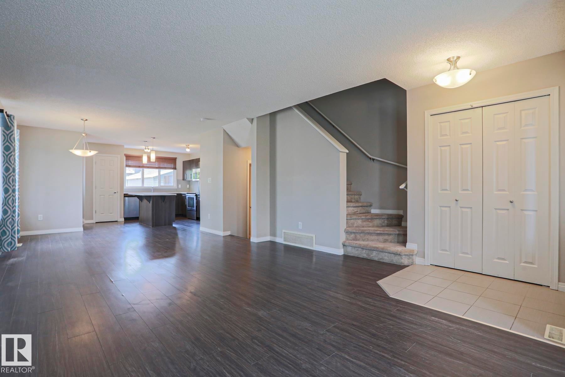 Open-concept living area with dark wood-finish flooring, featuring a kitchen island with pendant lighting, a carpeted staircase with a dark handrail, and a tiled entryway with double bifold doors - 489 Orchards Boulevard Sw, Edmonton, AB - Indoor Photo Showing Living Room