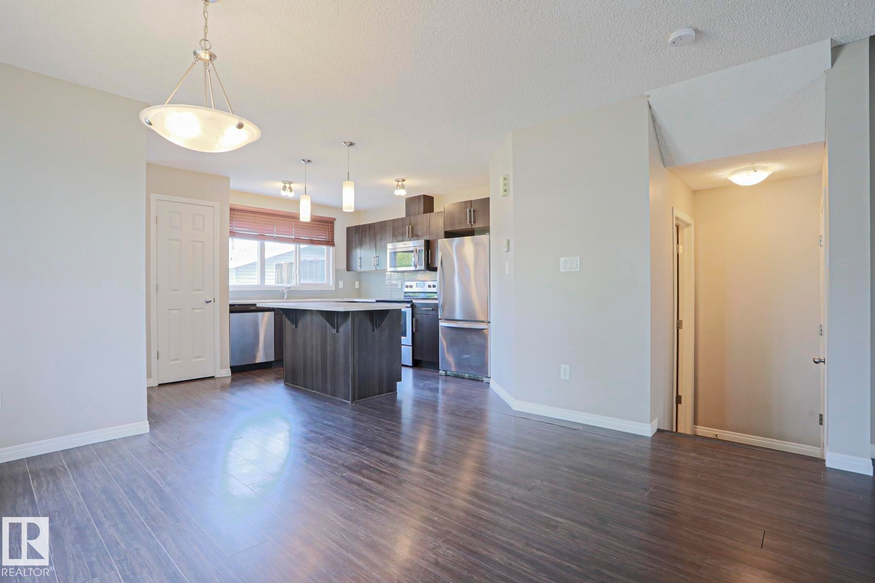 Open-concept living space featuring wood-finish flooring, a contemporary kitchen, stainless steel appliances, dark wood-finish cabinetry, and a central island with a light-colored countertop - 489 Orchards Boulevard Sw, Edmonton, AB - Indoor Photo Showing Other Room