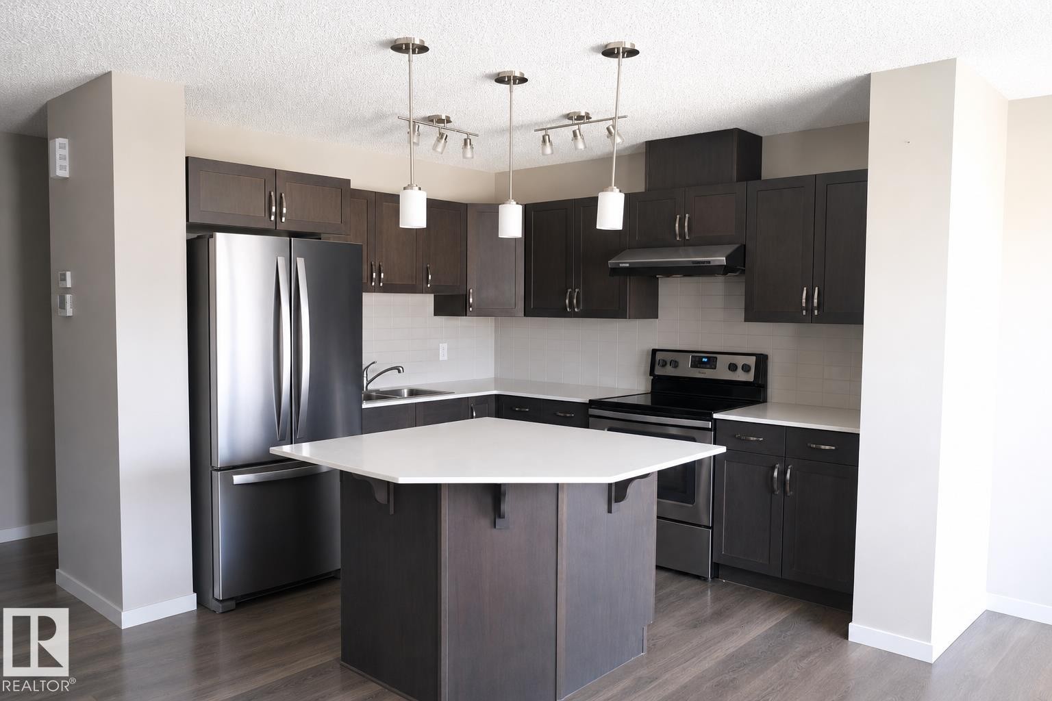 Modern kitchen featuring dark wood-finish cabinetry, stainless steel appliances, a white subway tile backsplash, and a hexagonal island with a light-colored countertop - 17263 9 Avenue Sw, Edmonton, AB