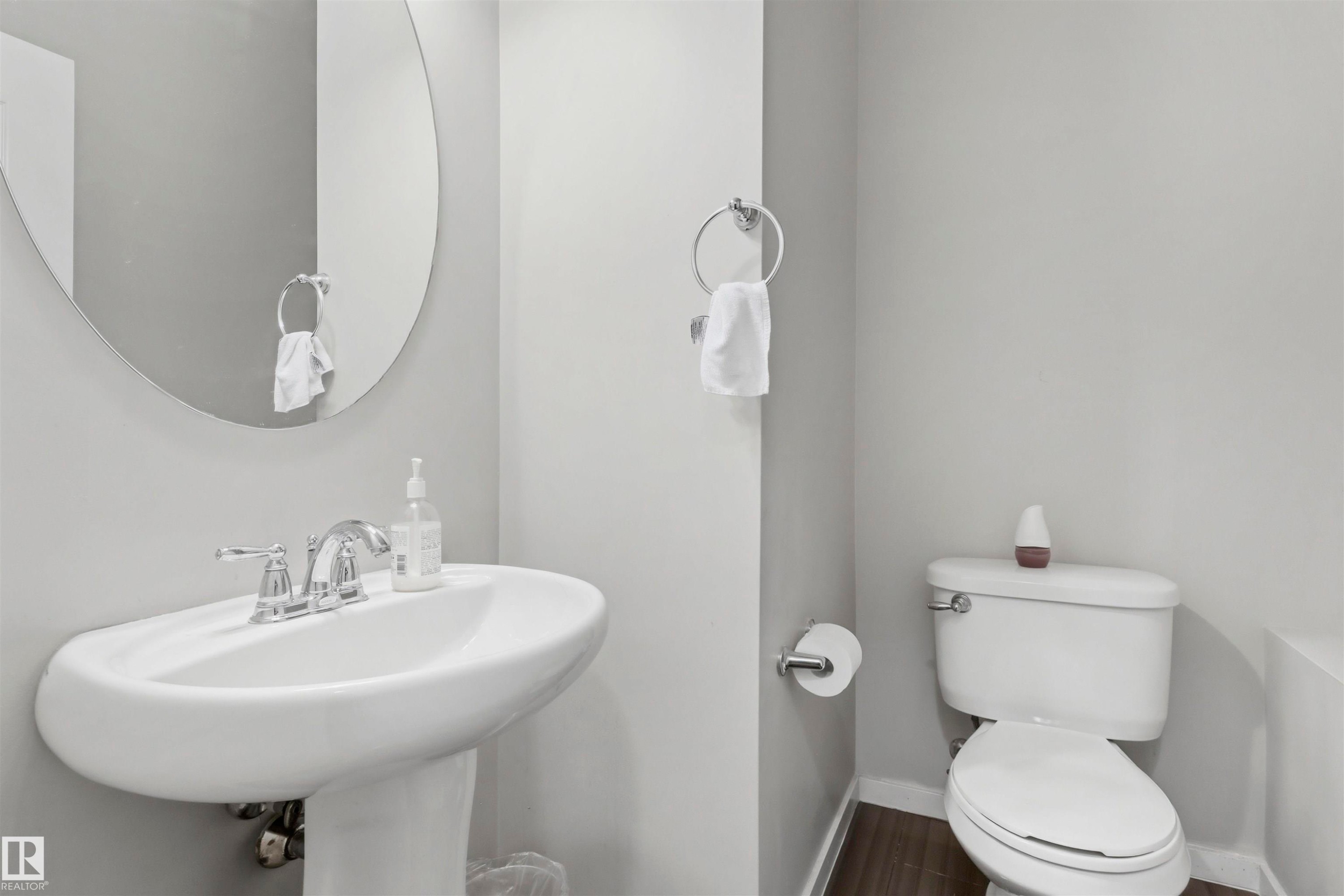 Pedestal sink with chrome faucet, oval mirror, white toilet, and wood-finish flooring - 1528 35 Avenue Nw, Edmonton, AB - Indoor Photo Showing Bathroom