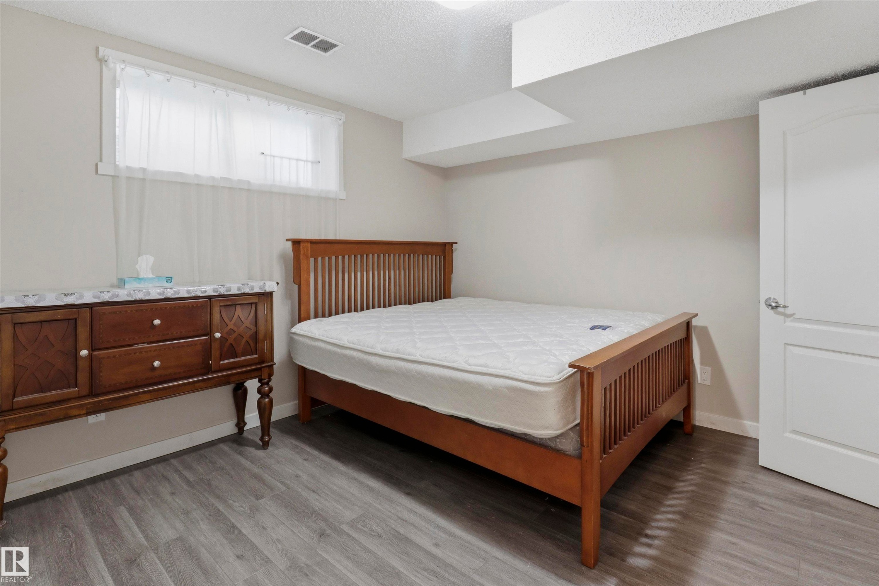 Bright room featuring wood-finish flooring, a window with sheer curtains, a white panel door, and neutral wall paint - 1528 35 Avenue Nw, Edmonton, AB - Indoor Photo Showing Bedroom