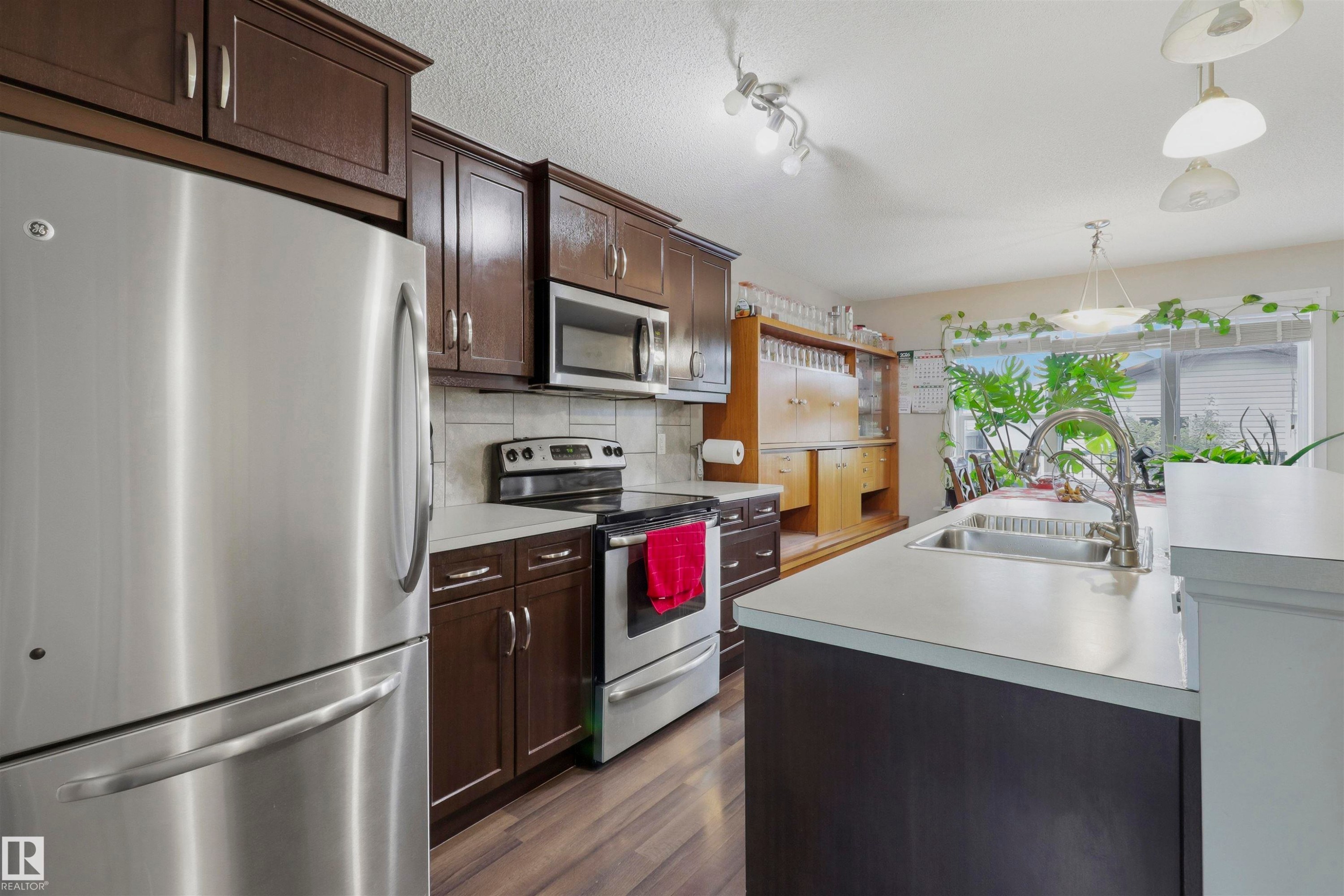Kitchen featuring dark wood cabinetry, stainless steel appliances, a tile backsplash, light-toned countertops, and wood-finish flooring - 1528 35 Avenue Nw, Edmonton, AB - Indoor Photo Showing Kitchen With Double Sink