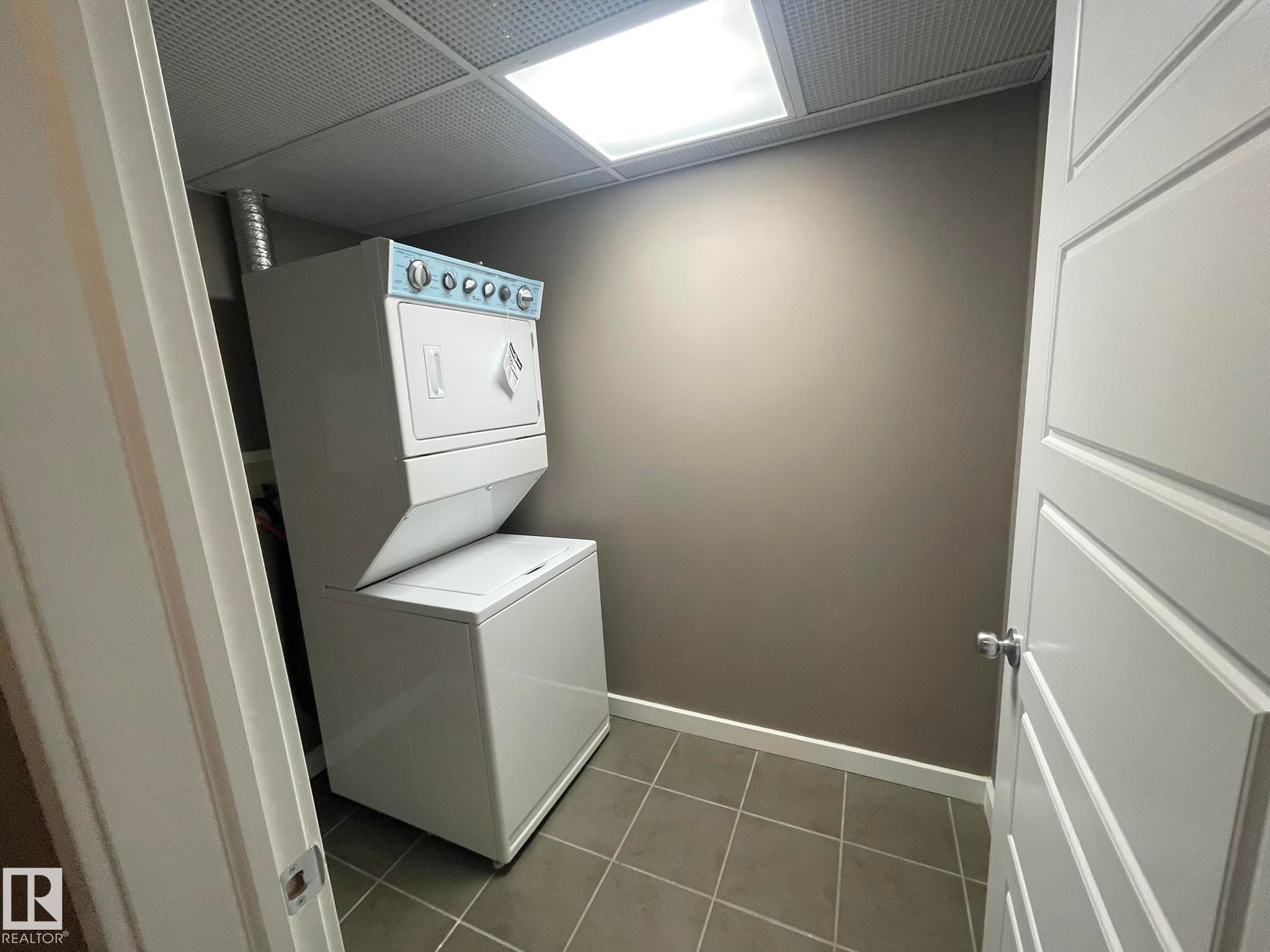 Dedicated laundry area featuring a stacked washer and dryer, square tile flooring, a drop ceiling with integrated lighting, and neutral wall paint - #504 10518 113 St Nw, Edmonton, AB - Indoor Photo Showing Laundry Room