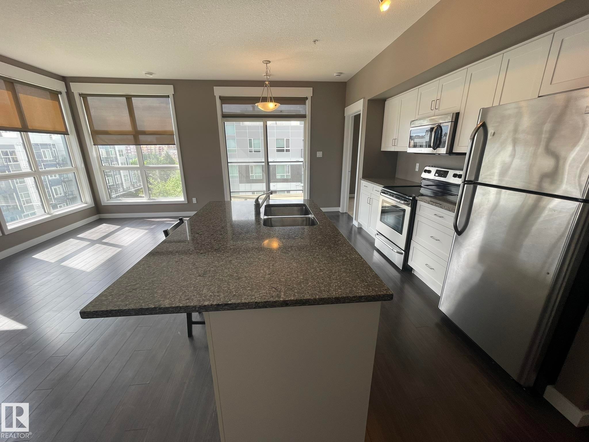 Open-concept kitchen featuring an island with a double basin sink, white cabinetry, stainless steel appliances, and dark wood-finish flooring - #504 10518 113 St Nw, Edmonton, AB - Indoor Photo Showing Kitchen With Stainless Steel Kitchen