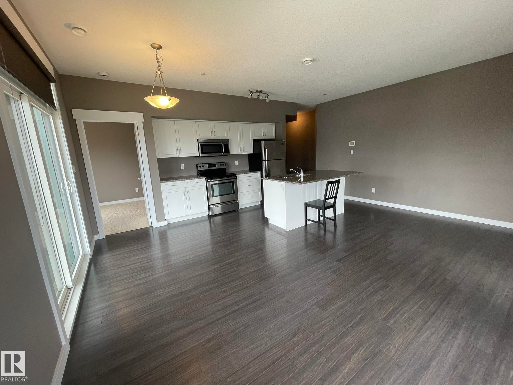 Open-concept living space featuring wood-finish flooring, a kitchen island with seating, stainless steel appliances, white cabinetry, and sliding glass patio doors - #504 10518 113 St Nw, Edmonton, AB - Indoor Photo Showing Other Room
