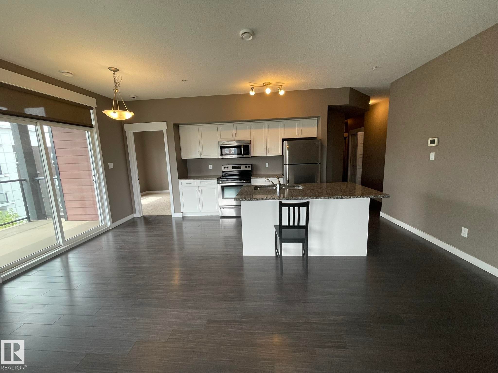 Open-concept living area featuring dark wood-finish flooring, a kitchen island with a stone-look countertop, white cabinetry, and stainless steel appliances - #504 10518 113 St Nw, Edmonton, AB - Indoor Photo Showing Kitchen