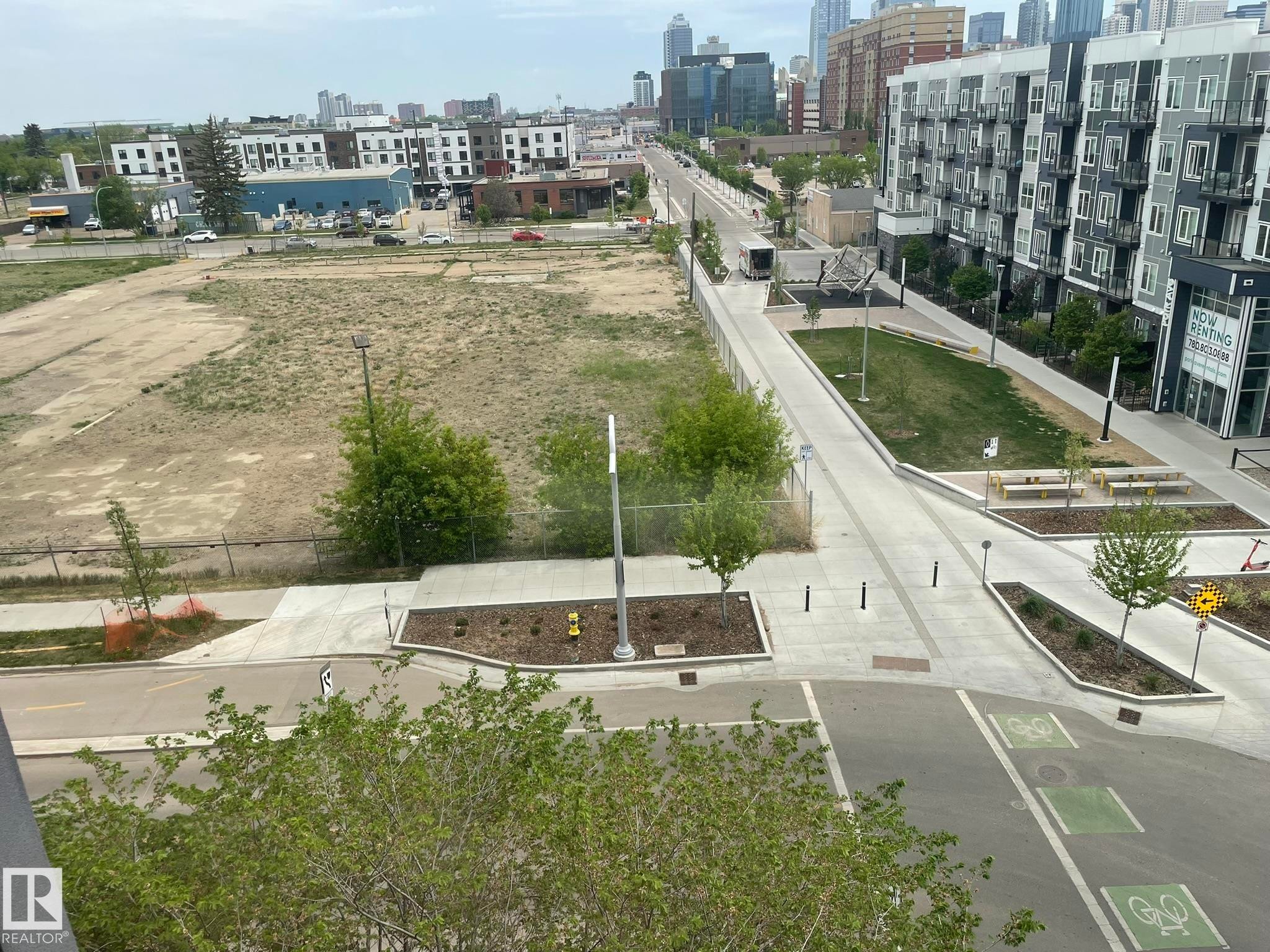 Urban landscape featuring a modern apartment building with balconies, extensive paved pathways, green spaces with planted trees, and bicycle lane markings on the street - #504 10518 113 St Nw, Edmonton, AB - Outdoor With View