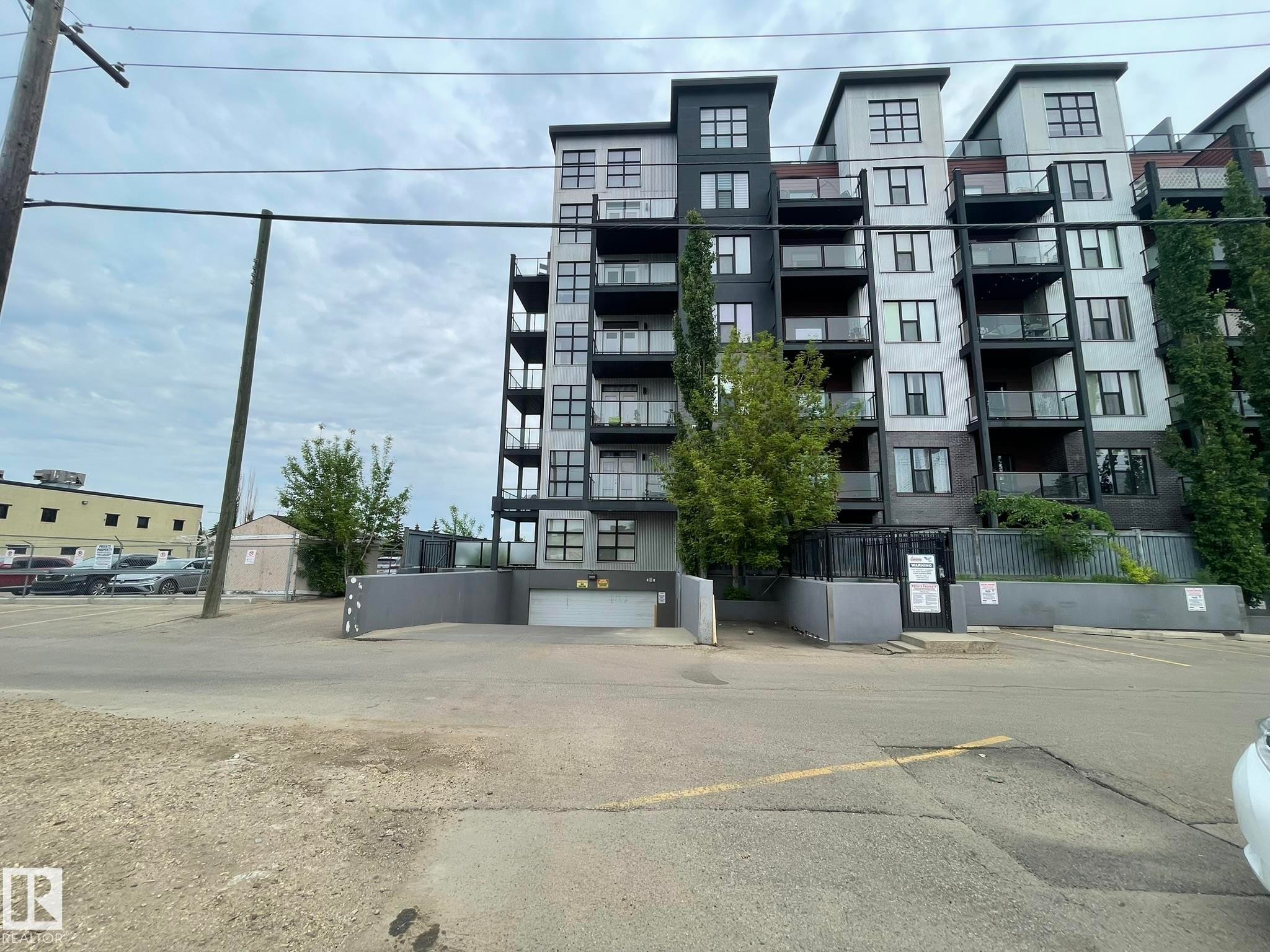 Contemporary multi-story building featuring a facade of light and dark paneling, individual balconies with glass railings, and a ground-level entrance to an underground parking facility - #504 10518 113 St Nw, Edmonton, AB - Outdoor With Balcony With Facade