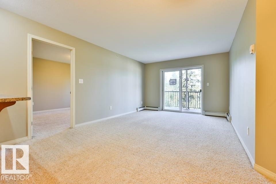 Spacious room featuring neutral carpeting, light-toned walls, and a sliding glass door providing access to an outdoor balcony with a railing - #406 4903 47 Av, Stony Plain, AB - Indoor Photo Showing Other Room