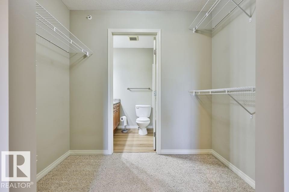 Carpeted closet with white wire shelving, providing access to a bathroom with wood-finish flooring, a vanity with wood cabinetry, a light-toned countertop, and a white toilet - #406 4903 47 Av, Stony Plain, AB - Indoor With Storage