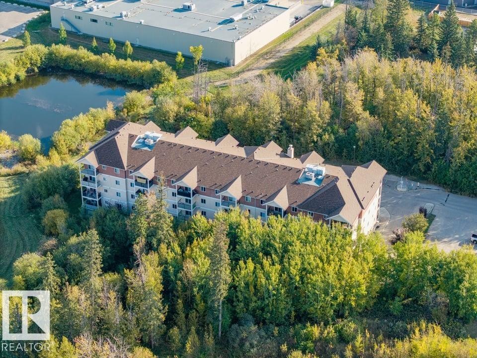 Multi-story building surrounded by mature trees, featuring a brown shingle roof, multiple balconies, and an adjacent parking area - #406 4903 47 Av, Stony Plain, AB - Outdoor With View