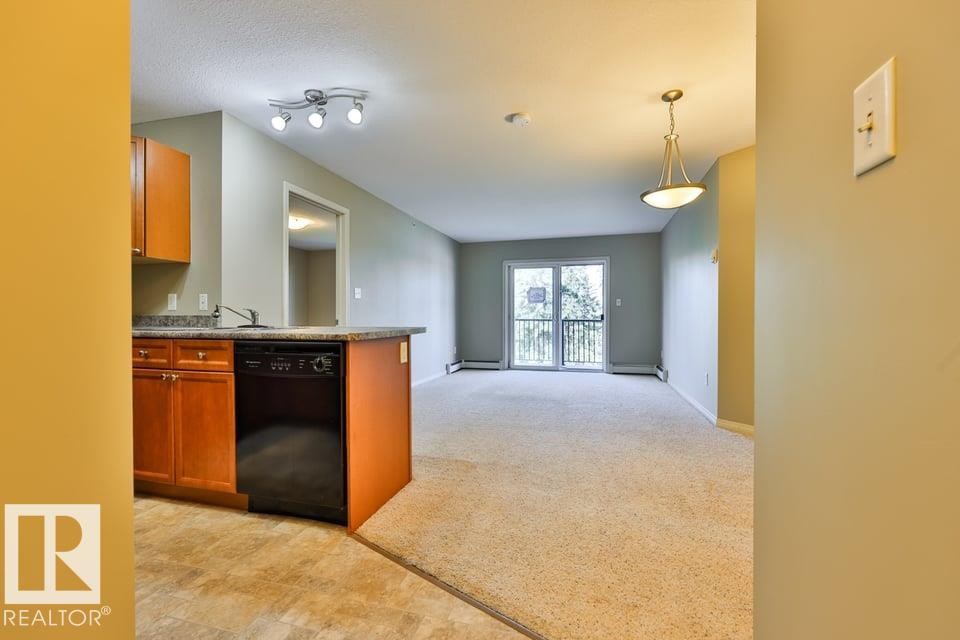 Kitchen with wood cabinetry and dark countertops, integrated with a carpeted living area featuring a sliding glass door to an exterior balcony - #406 4903 47 Av, Stony Plain, AB - Indoor