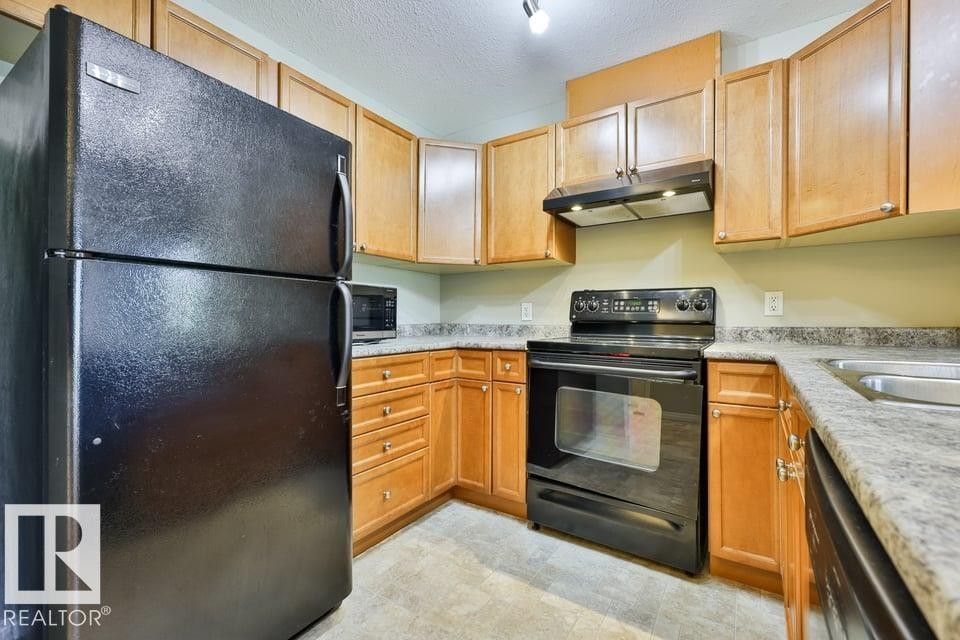 Kitchen featuring wood cabinetry, black appliances, a double-basin stainless steel sink, and light-toned countertops - #406 4903 47 Av, Stony Plain, AB - Indoor Photo Showing Kitchen