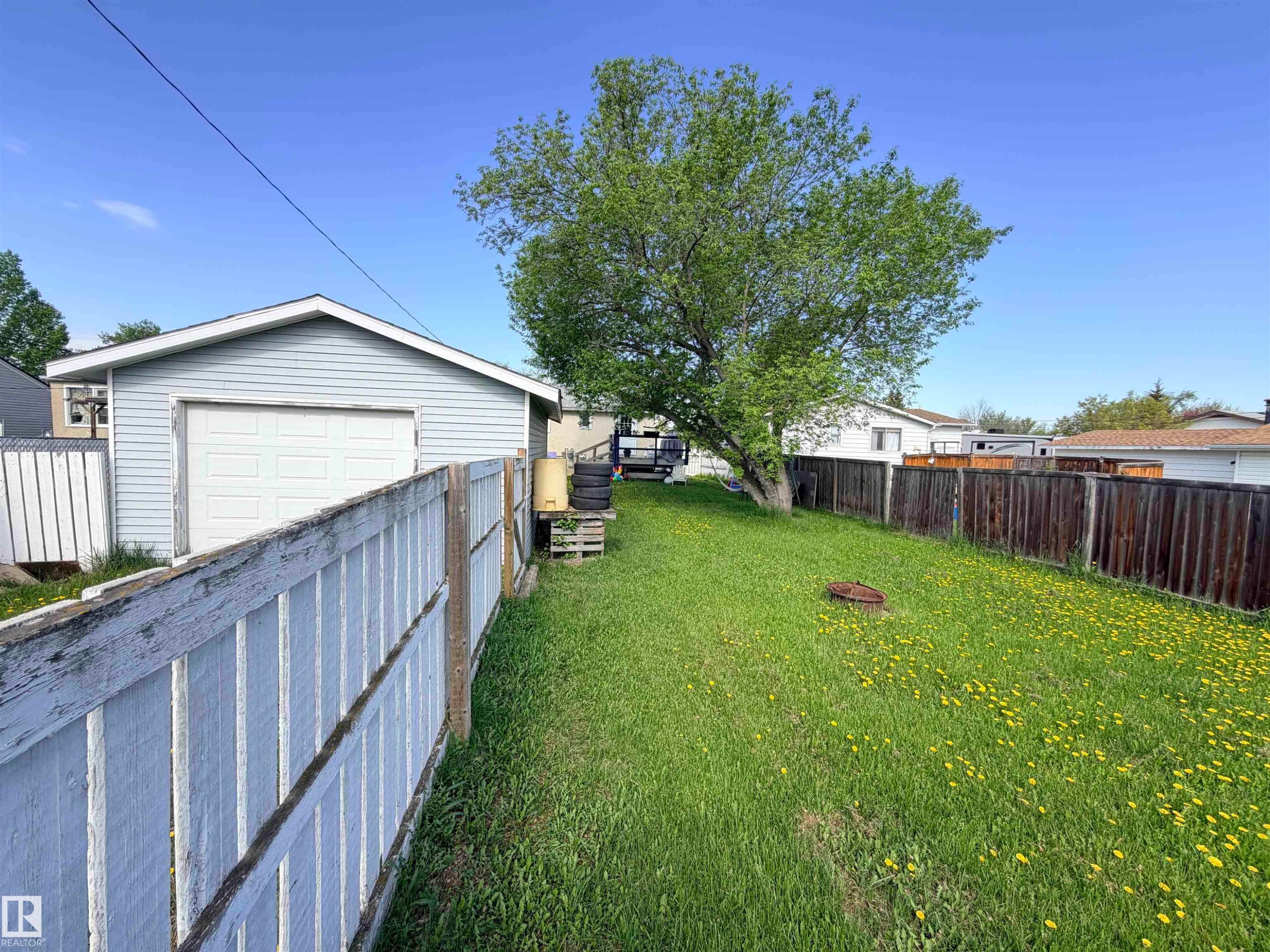 Expansive backyard featuring a mature deciduous tree, a detached garage with vinyl siding and a roll-up door, and a perimeter defined by both light-colored and dark-stained wooden fencing - 4823 49 Street, Clyde, AB - Outdoor