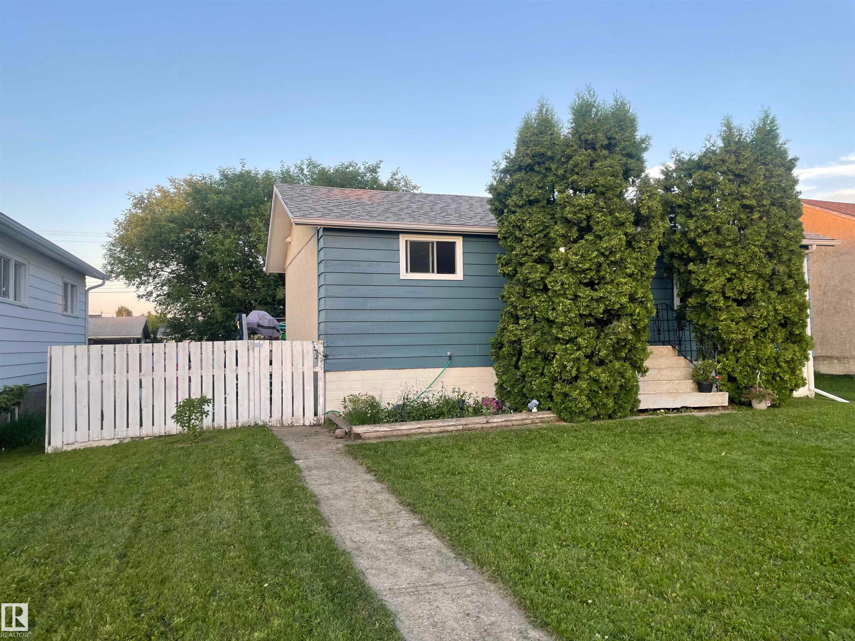 Gable roof home featuring horizontal siding, a concrete pathway, a white picket fence, and mature evergreen shrubs - 4823 49 Street, Clyde, AB - Outdoor
