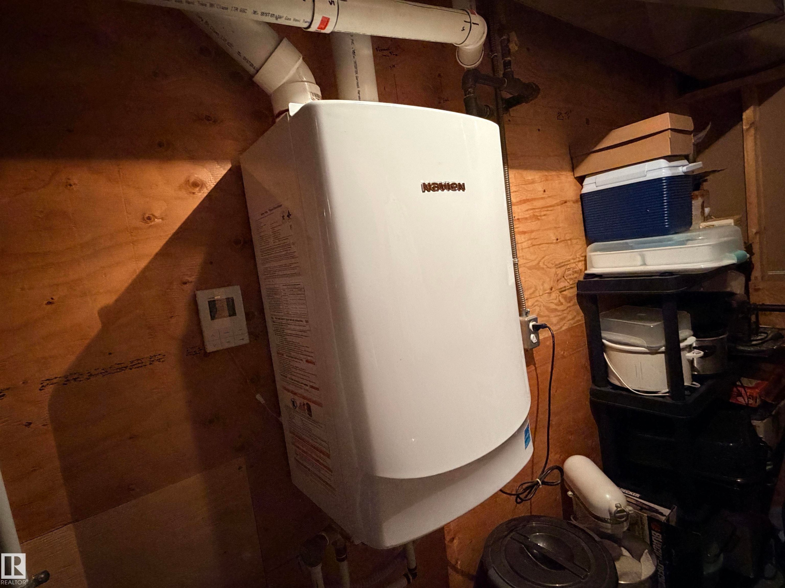 Wall-mounted water heater with visible PVC piping, set against a plywood wall - 4823 49 Street, Clyde, AB - Indoor Photo Showing Other Room