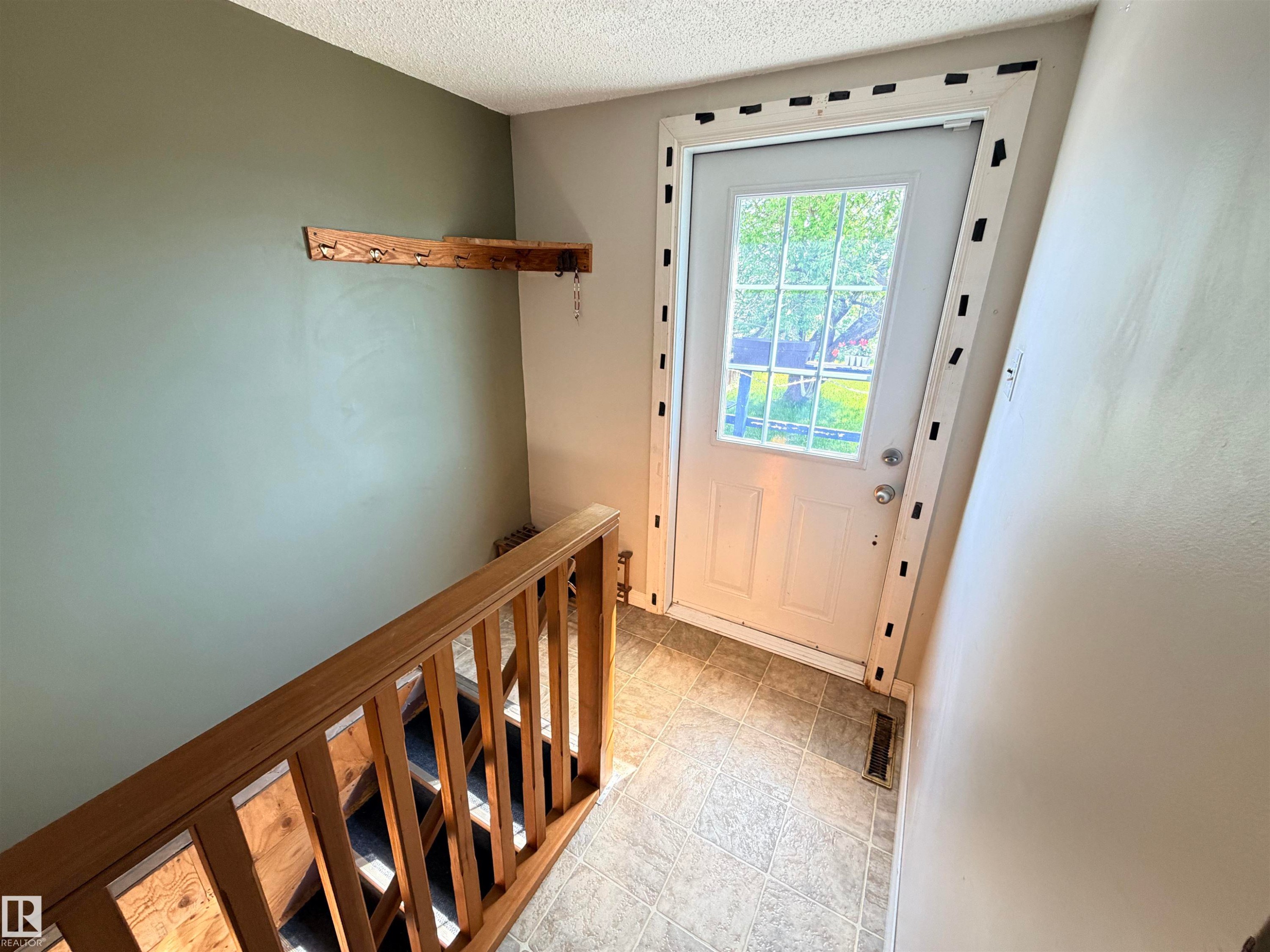 Entryway featuring a paneled door with grid-patterned window, tiled flooring, wood-finish stair railing, and a rustic wood-finish coat rack - 4823 49 Street, Clyde, AB - Indoor Photo Showing Other Room