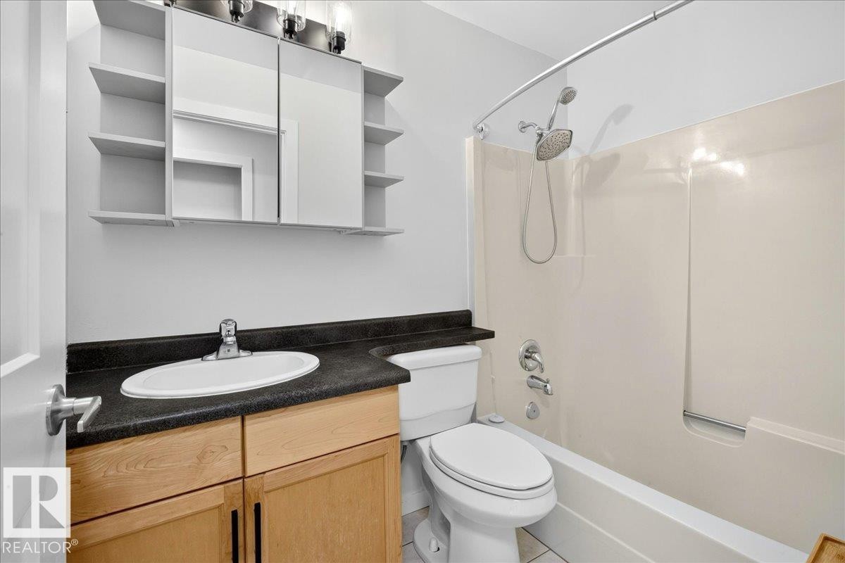 Full bathroom featuring a light wood-finish vanity with dark countertop, an oval undermount sink, a shower-tub combination with a curved shower rod, and a mirrored medicine cabinet with integrated shelving - 4212 32 Avenue Nw, Edmonton, AB - Indoor Photo Showing Bathroom