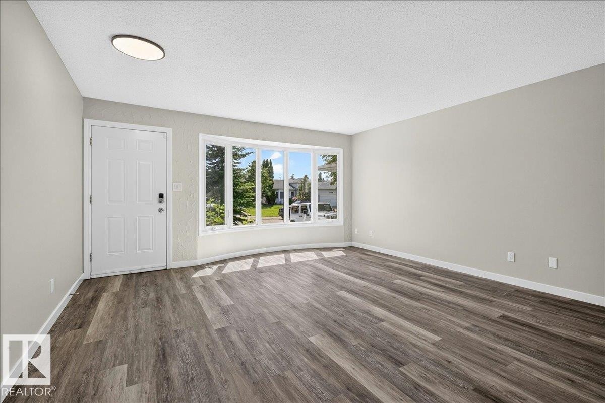 Spacious room featuring wood-finish flooring, a contemporary ceiling light fixture, a paneled entry door with smart lock, and a large bay window - 4212 32 Avenue Nw, Edmonton, AB - Indoor
