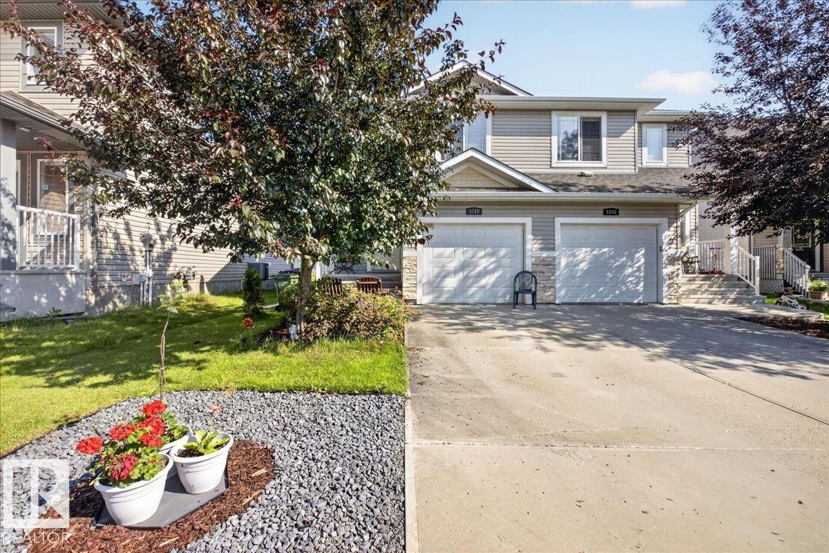 Exterior featuring light-colored siding with stone accents, a two-car garage with individual doors, a concrete driveway, a front lawn with mature trees, and a decorative gravel landscaping element - 5110 168A Avenue Nw, Edmonton, AB - Outdoor
