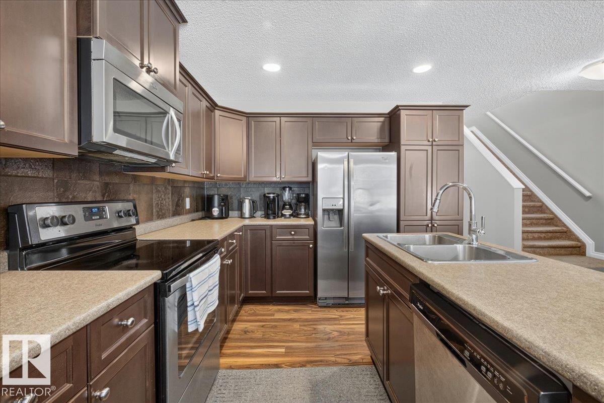 Modern kitchen featuring rich cabinetry, speckled countertops, stainless steel appliances, a tile backsplash, and wood-finish flooring - 5110 168A Avenue Nw, Edmonton, AB - Indoor Photo Showing Kitchen With Double Sink