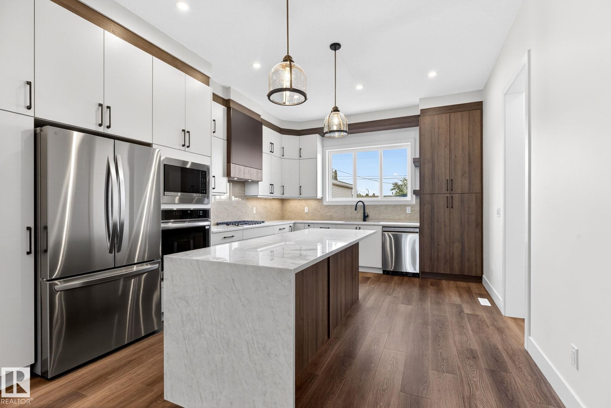 Contemporary kitchen featuring a large waterfall island with a stone-finish countertop, dark wood-finish cabinetry, stainless steel appliances, pendant lighting, and wood-finish flooring - 5308 103A Avenue, Edmonton, AB - Indoor Photo Showing Kitchen With Upgraded Kitchen