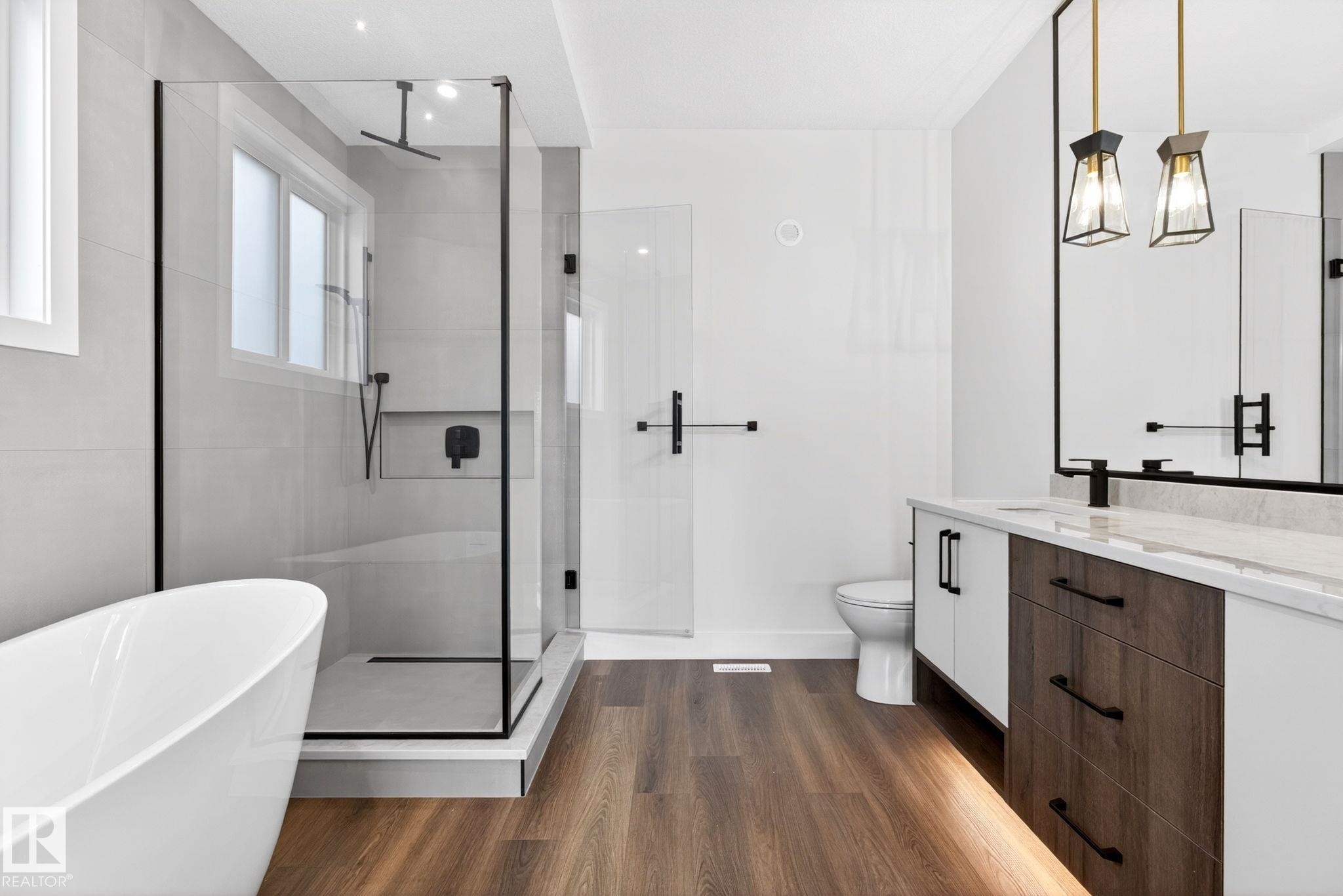 Contemporary bathroom featuring a freestanding soaking tub, a frameless glass shower enclosure with matte black fixtures, wood-finish flooring, a dual-sink vanity with a light-colored countertop, and modern pendant lighting - 5308 103A Avenue, Edmonton, AB - Indoor Photo Showing Bathroom