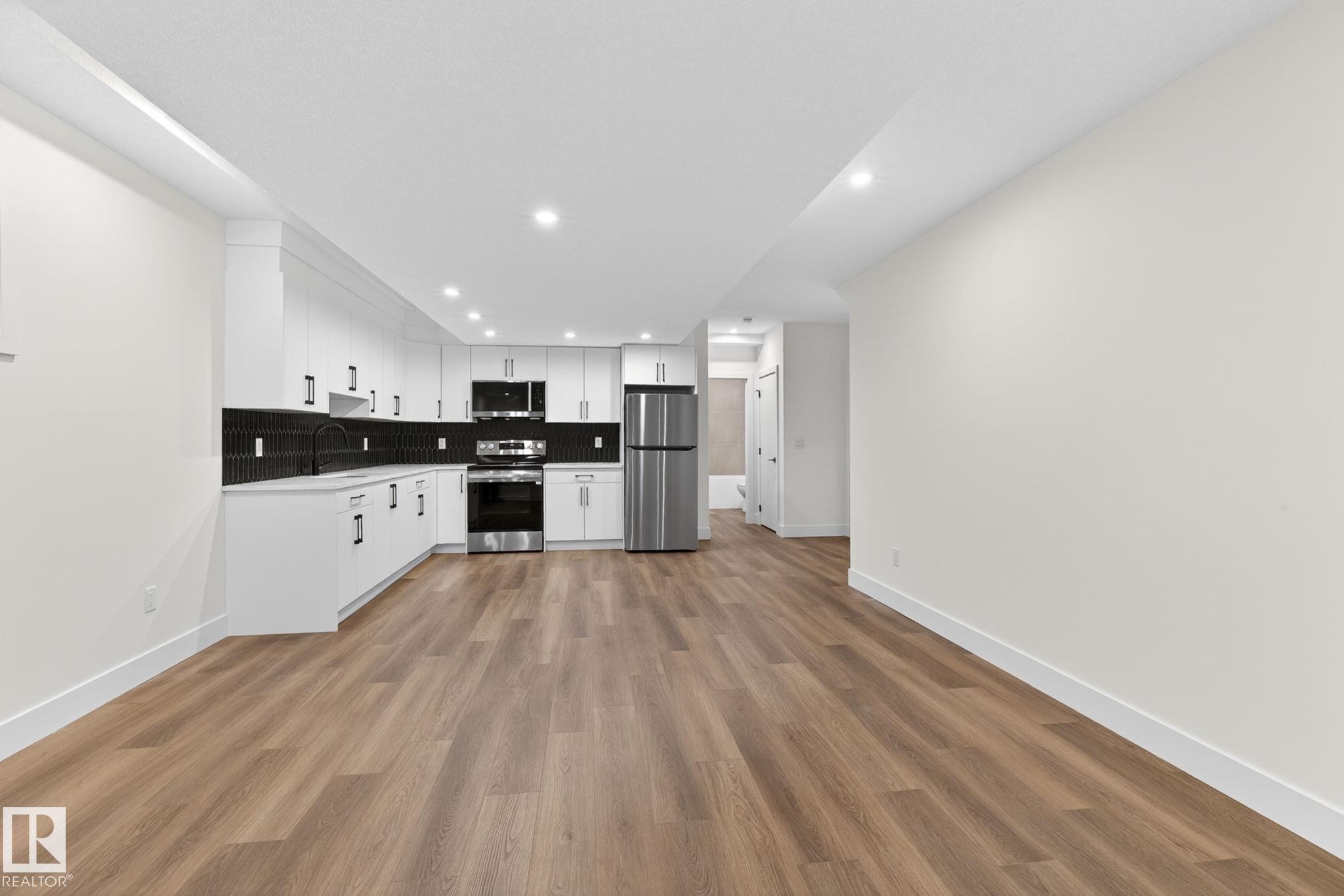 Open-concept living area featuring wood-finish flooring, recessed lighting, and a kitchen with white cabinetry, stainless steel appliances, and a black tile backsplash - 5308 103A Avenue, Edmonton, AB - Indoor Photo Showing Kitchen