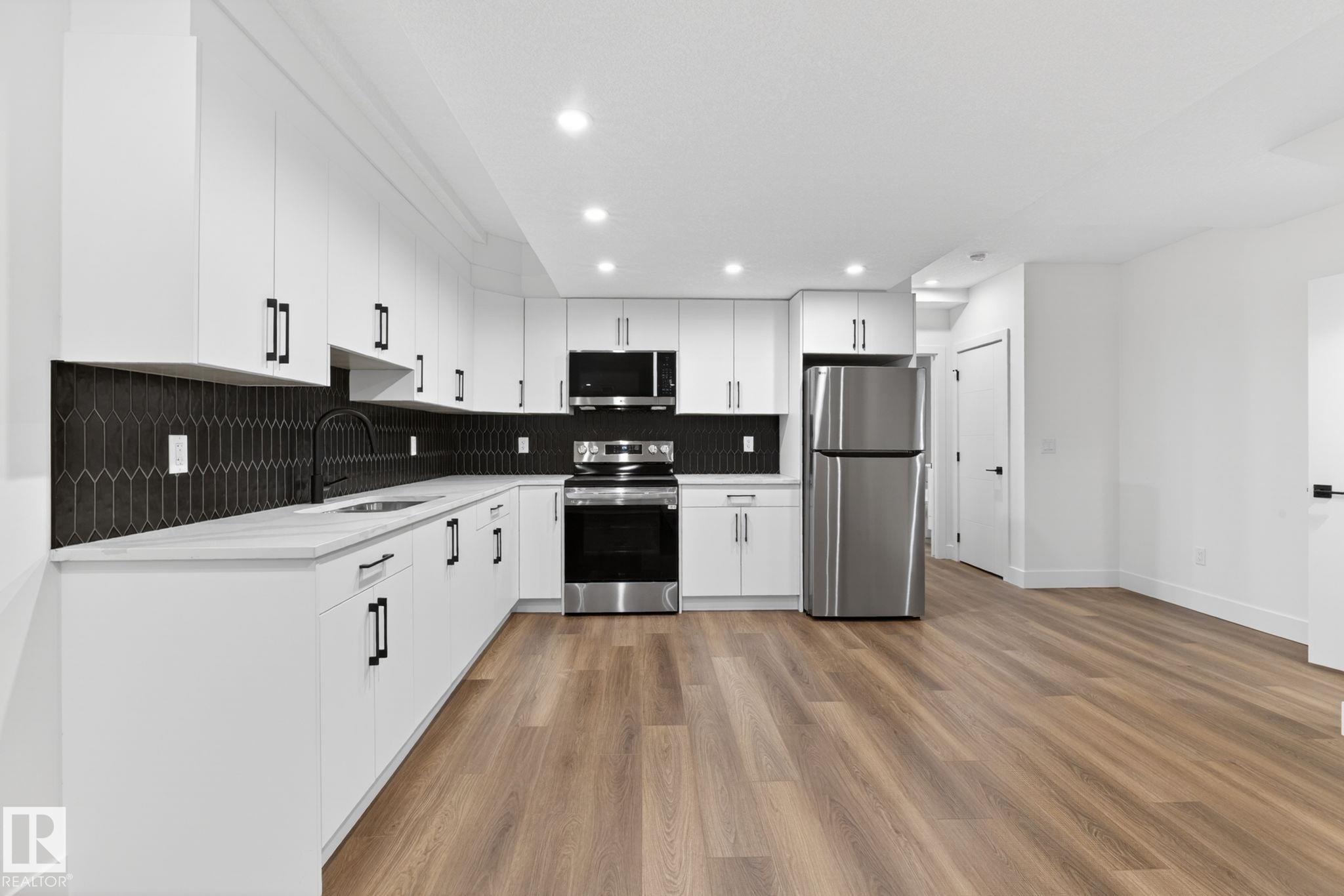 Modern kitchen featuring white cabinetry with matte black hardware, stainless steel appliances, a dark geometric tile backsplash, light countertops, and wood-finish flooring - 5308 103A Avenue, Edmonton, AB - Indoor Photo Showing Kitchen