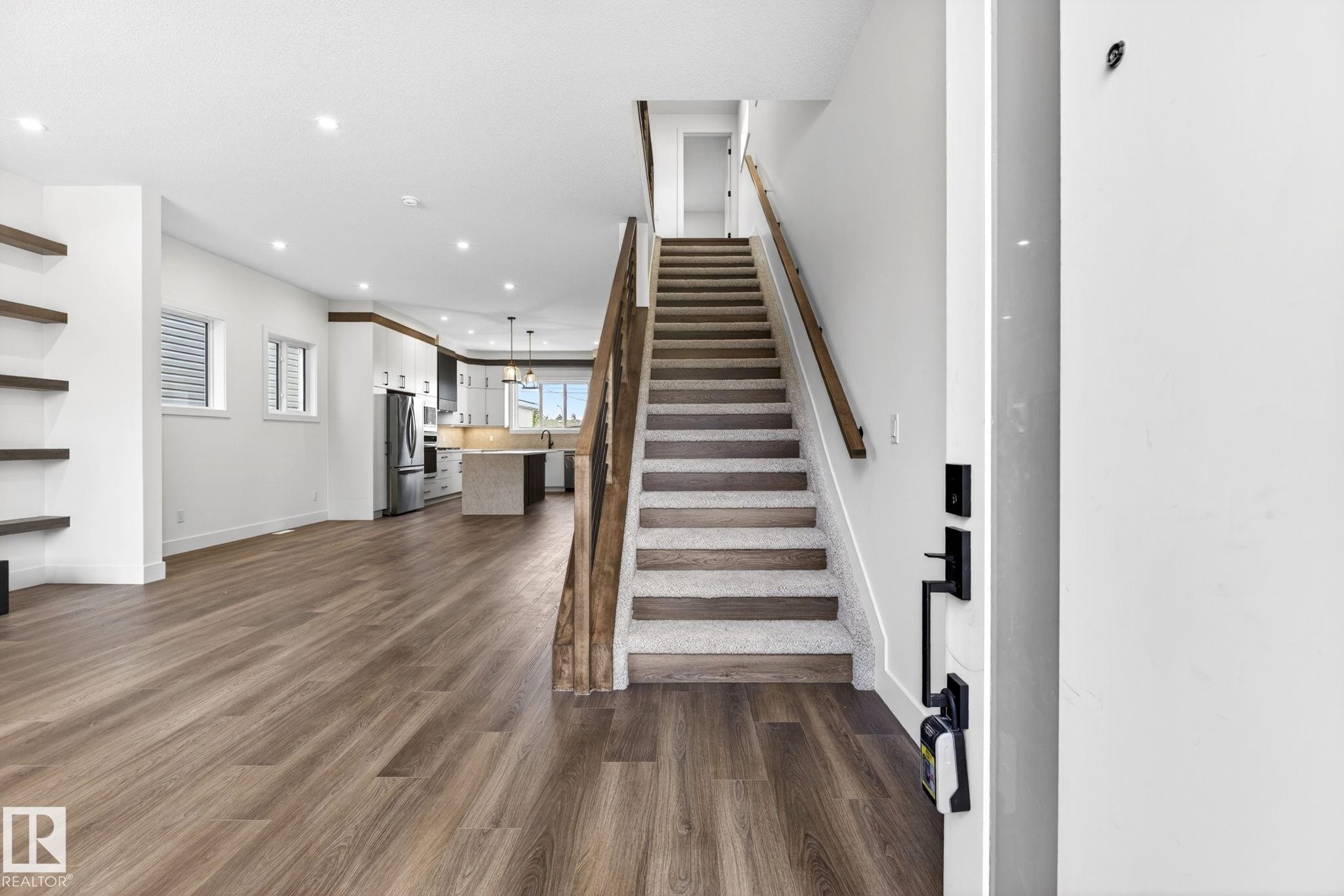 Open-concept living space featuring wood-finish flooring, recessed lighting, floating wall shelving, a carpeted staircase with wood railing, and a kitchen with an island - 5308 103A Avenue, Edmonton, AB - Indoor Photo Showing Other Room