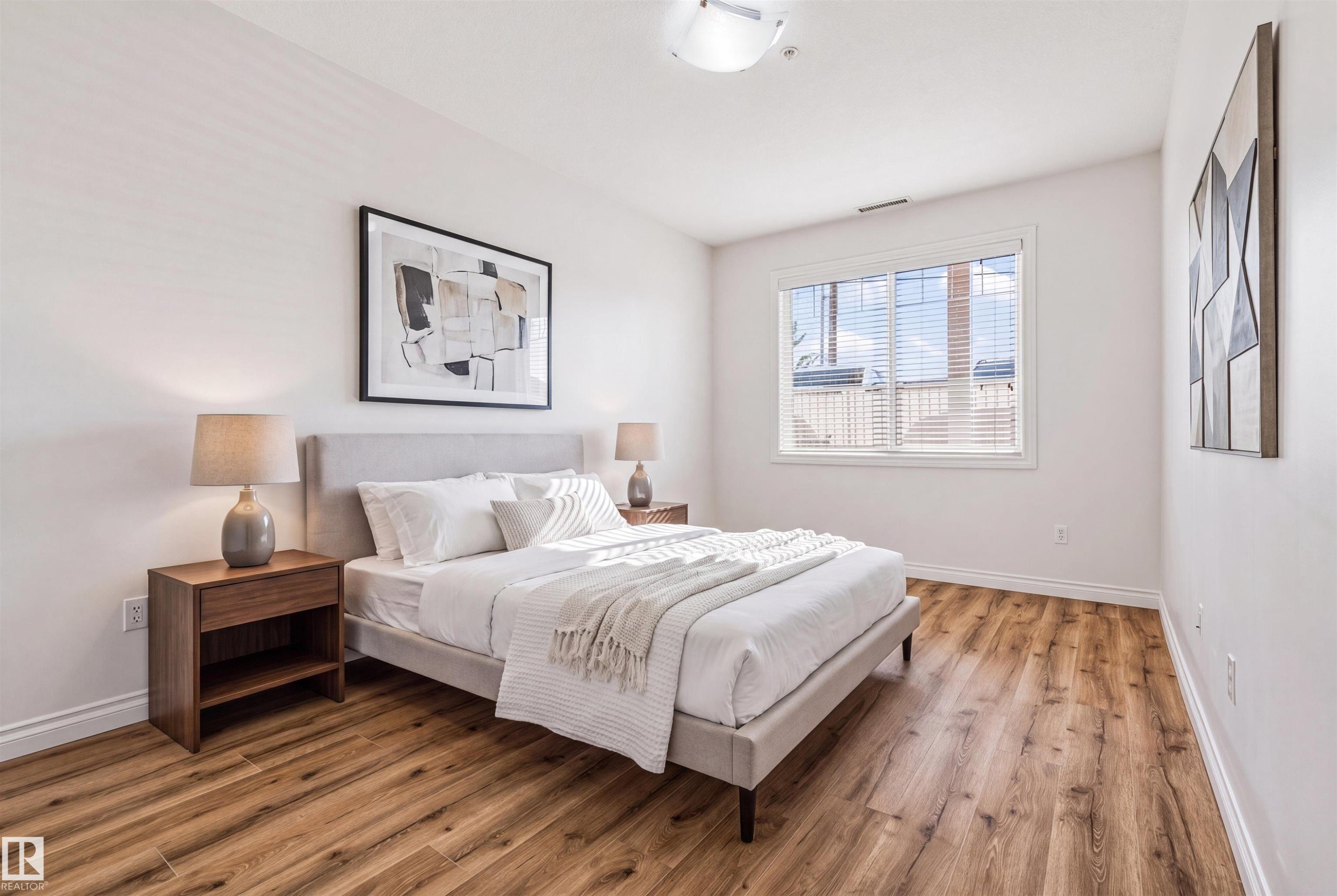 Bright primary bedroom featuring wood-finish flooring, white baseboards, and a large window with horizontal blinds - #110 5211 50 St, Stony Plain, AB - Indoor Photo Showing Bedroom