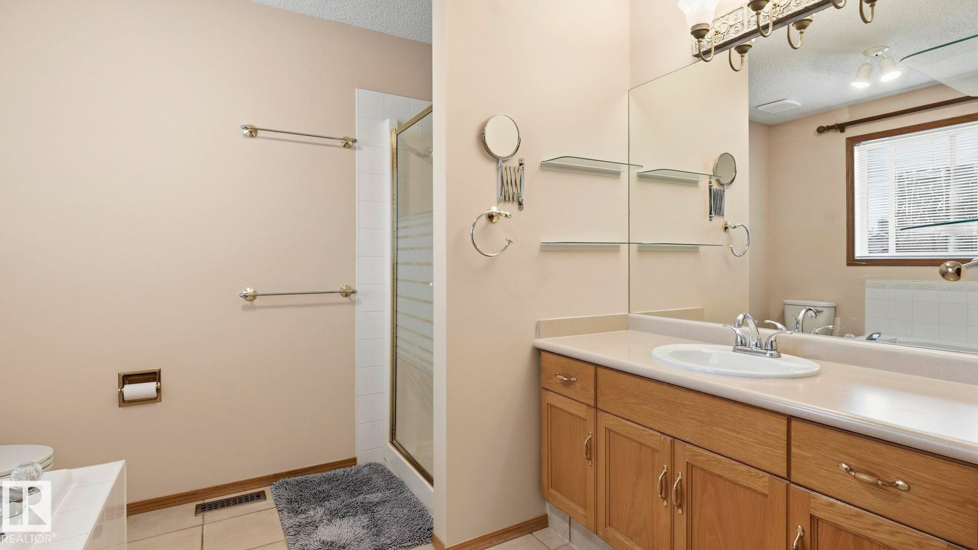 Bathroom featuring a glass-enclosed shower, light wood-finish vanity with ample storage, a white basin, a large mirror, and wall-mounted shelving - 125 Dechene Road Nw, Edmonton, AB - Indoor Photo Showing Bathroom