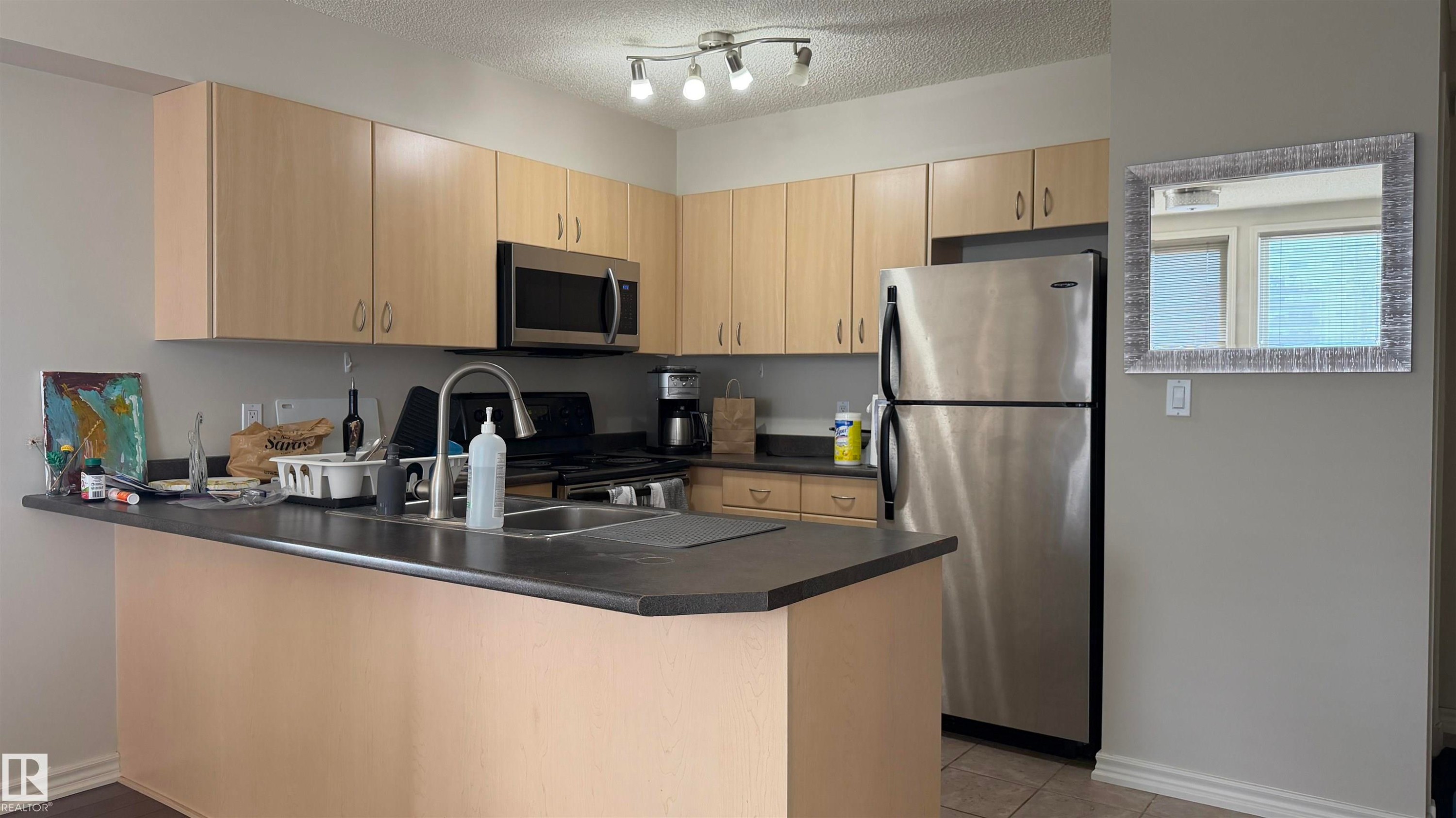 Kitchen featuring light wood-finish cabinetry, stainless steel appliances, and a peninsula with a dark countertop - #2401 10180 104 St Nw, Edmonton, AB - Indoor Photo Showing Kitchen With Double Sink
